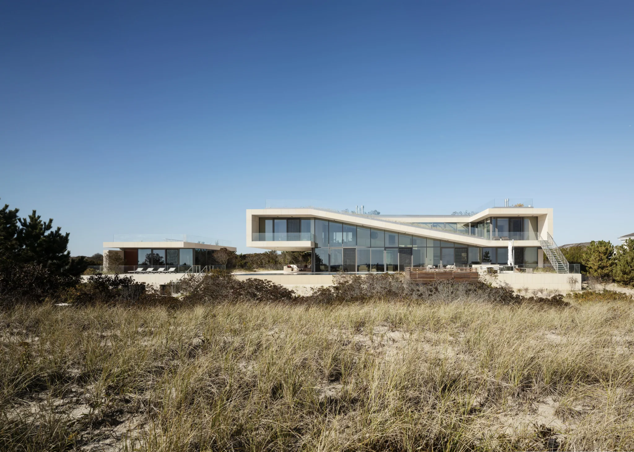 Custom oceanfront house with angular terraces and expansive glass walls set behind coastal dunes under a clear sky