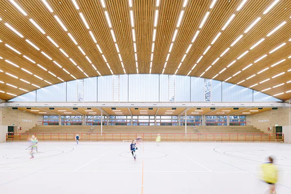 Interior of Böllenfalltorhalle sports hall in Darmstadt with wood slat ceiling, natural light, bleacher seating, and athletes playing indoors.