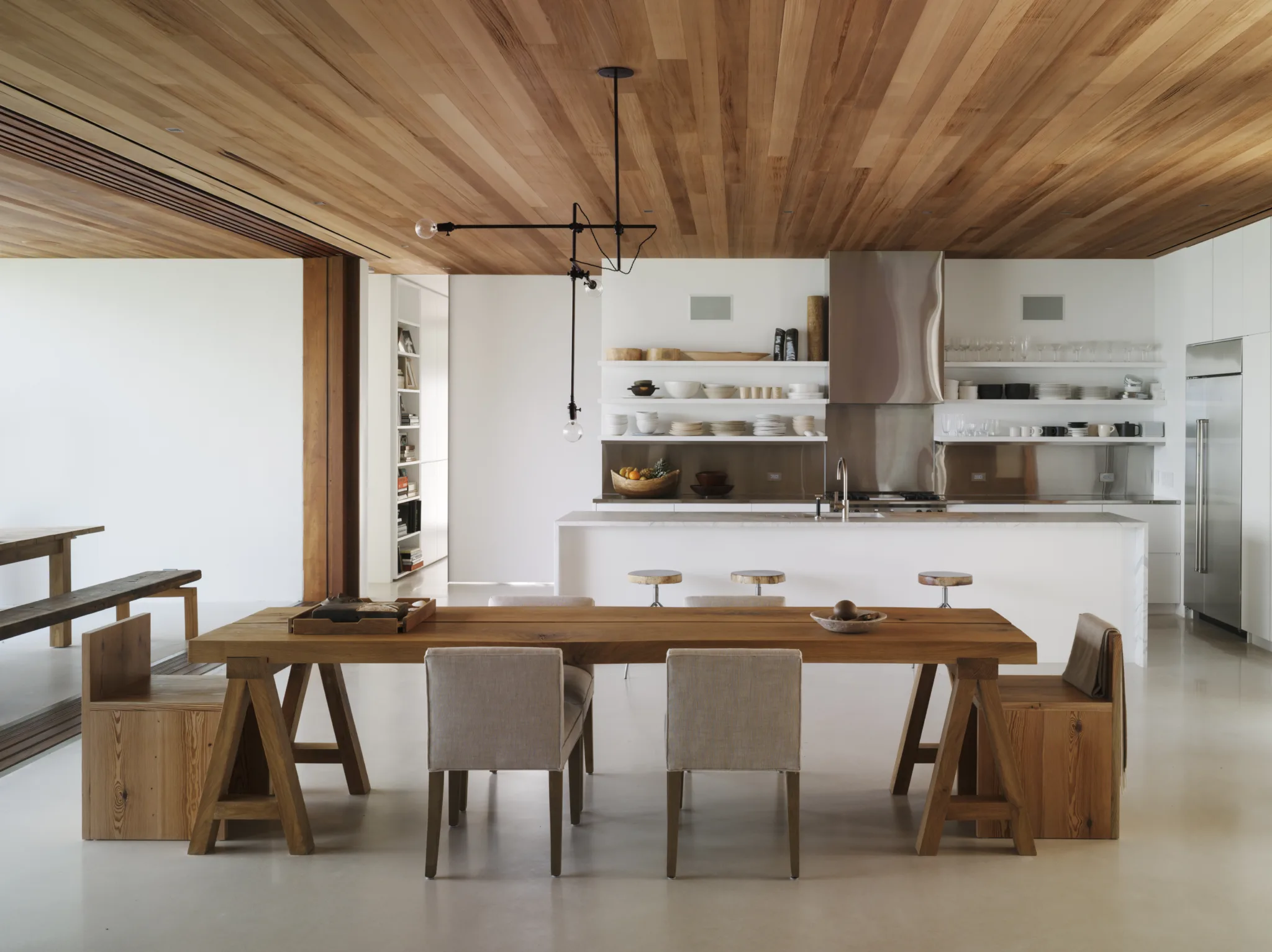 A modern indoor/outdoor dining room and kitchen with white concrete floors, white walls and teak ceiling.