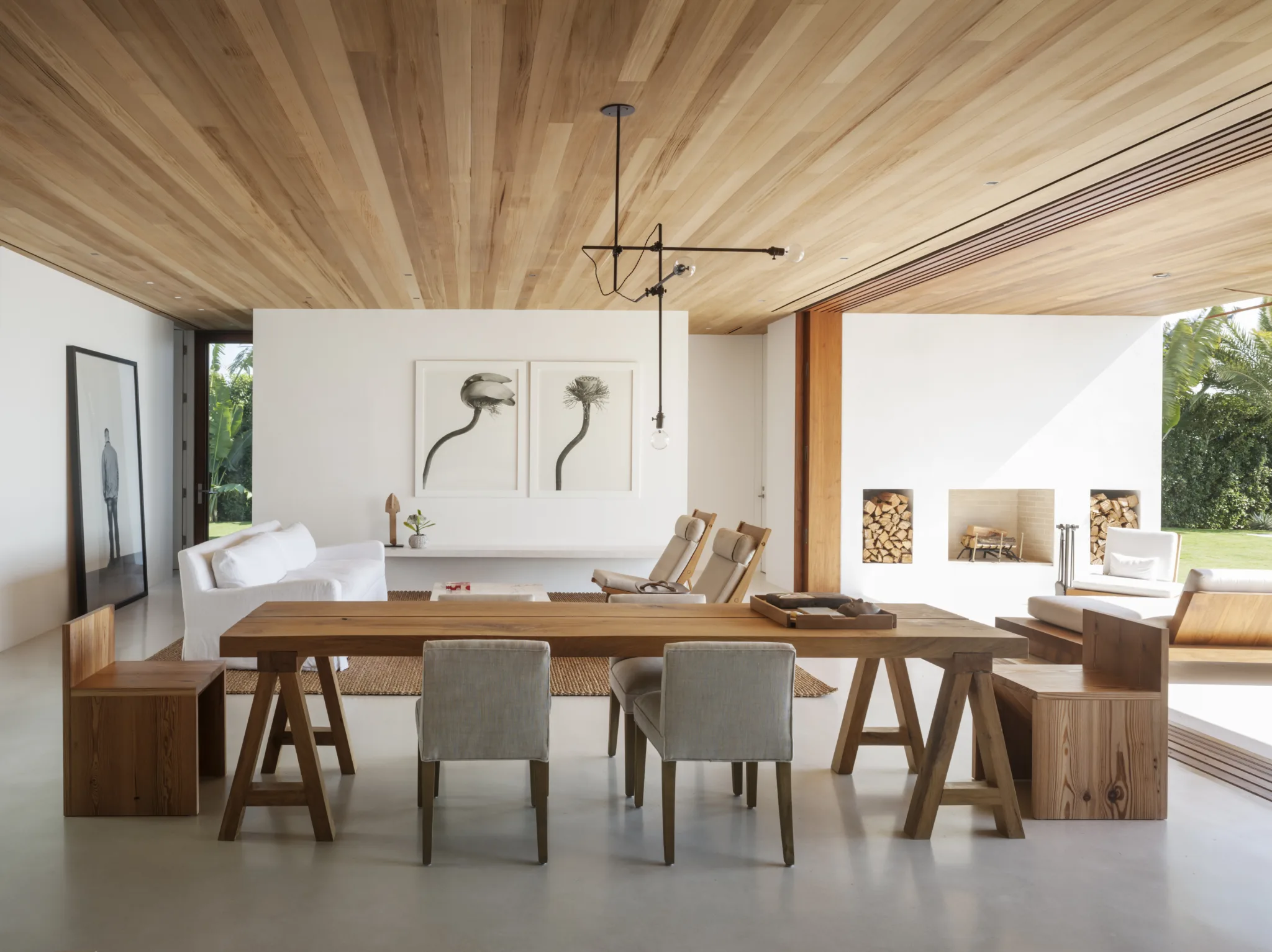 A modern indoor/outdoor dining room and living room with white concrete floors, white walls and teak ceiling. The doors to the waterfront pull completely out of view to show outdoor seating and fireplace wall.