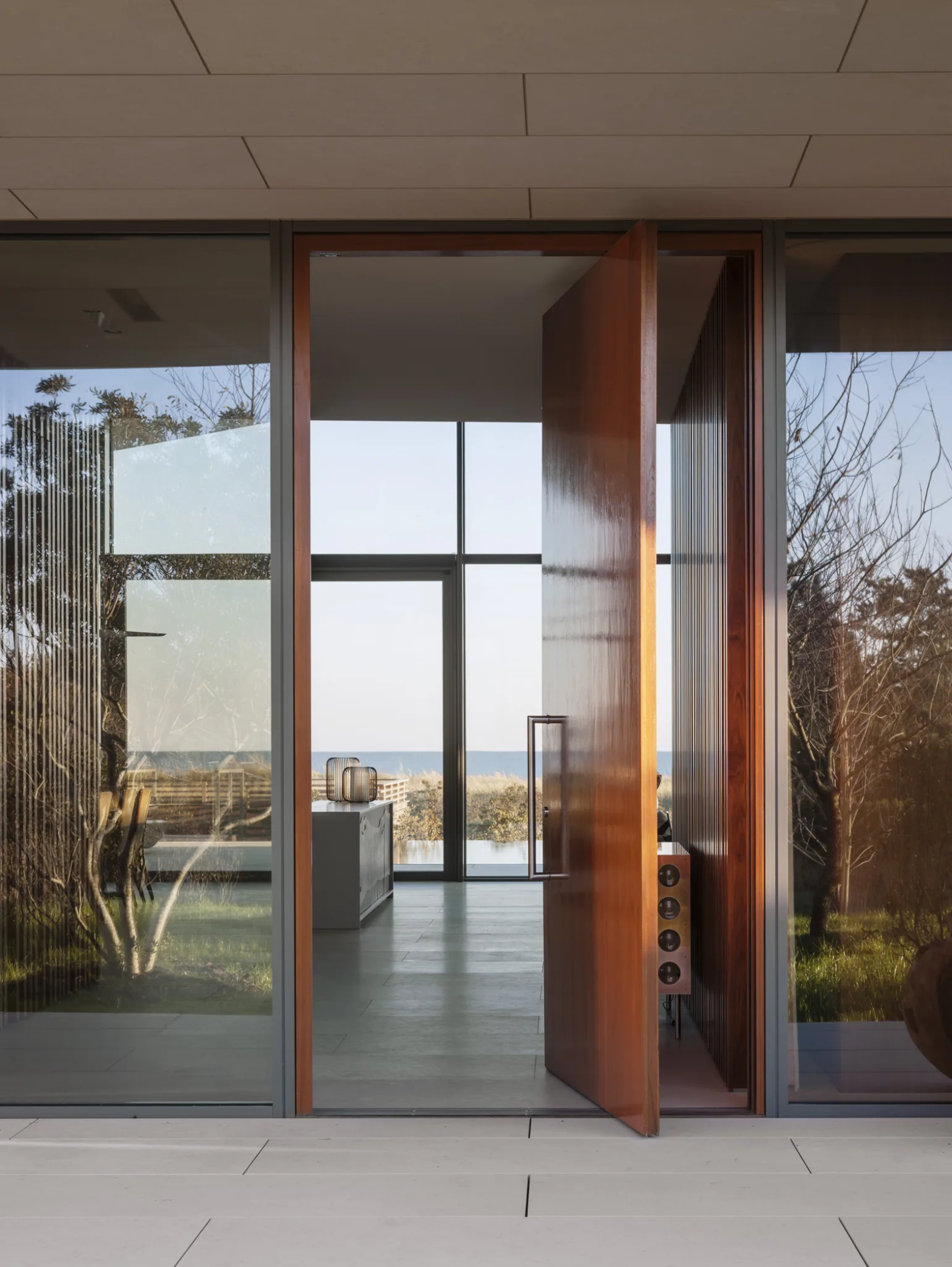 Modern beachfront entryway with pivoting wooden door, glass walls, and ocean views framed through the minimalist interior.