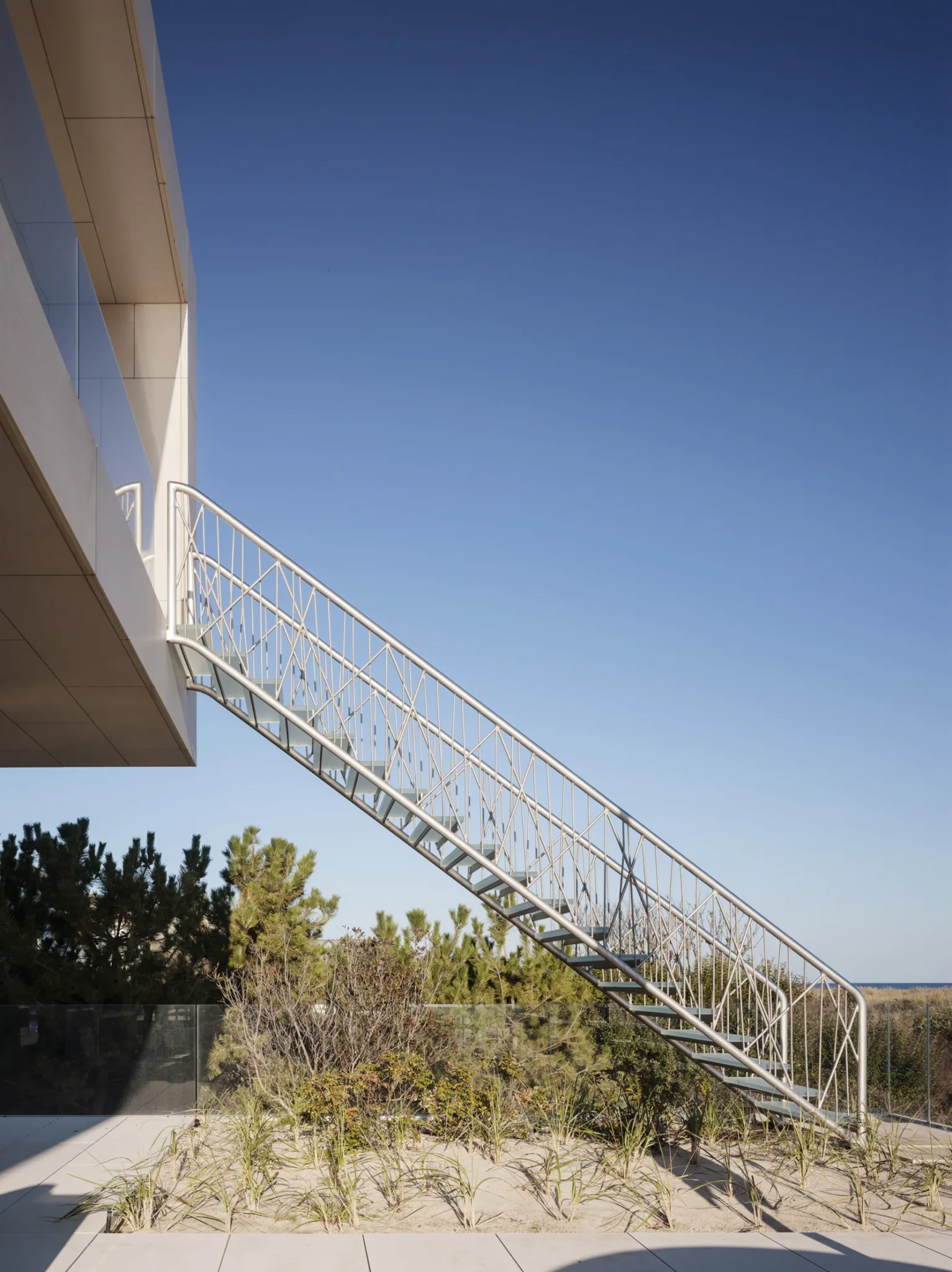Exterior staircase leading from upper terrace to beach landscape, showcasing geometric metalwork against a clear blue sky.