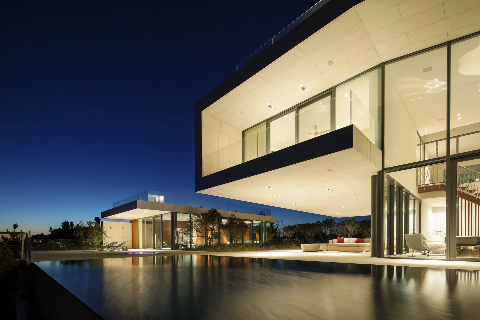 Modern Long Island beach house at dusk with cantilevered upper level, floor-to-ceiling glass, and pool reflecting the evening sky.