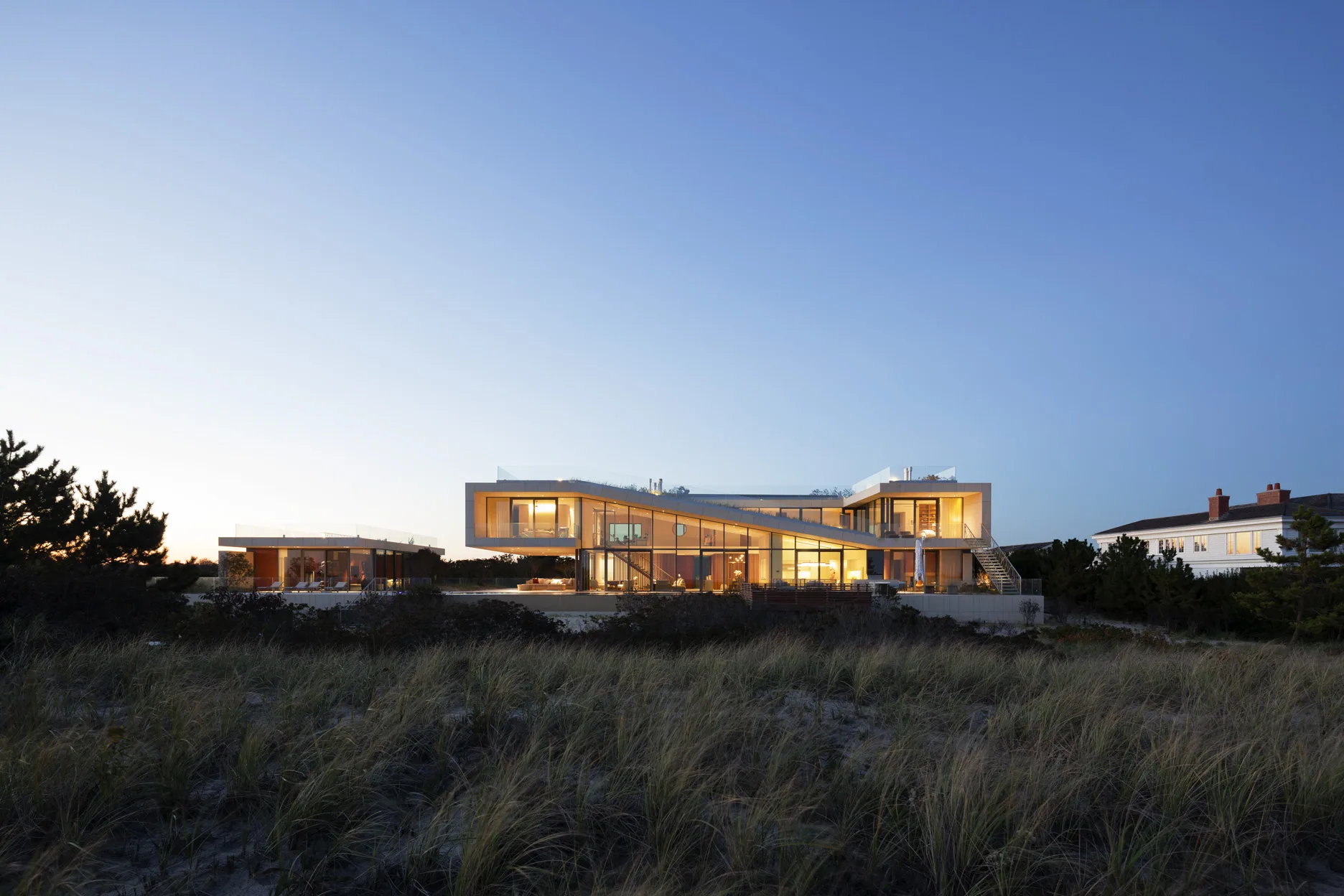Modern Long Island beach house with glowing glass façade and angular roofline set against the twilight sky and coastal landscape.