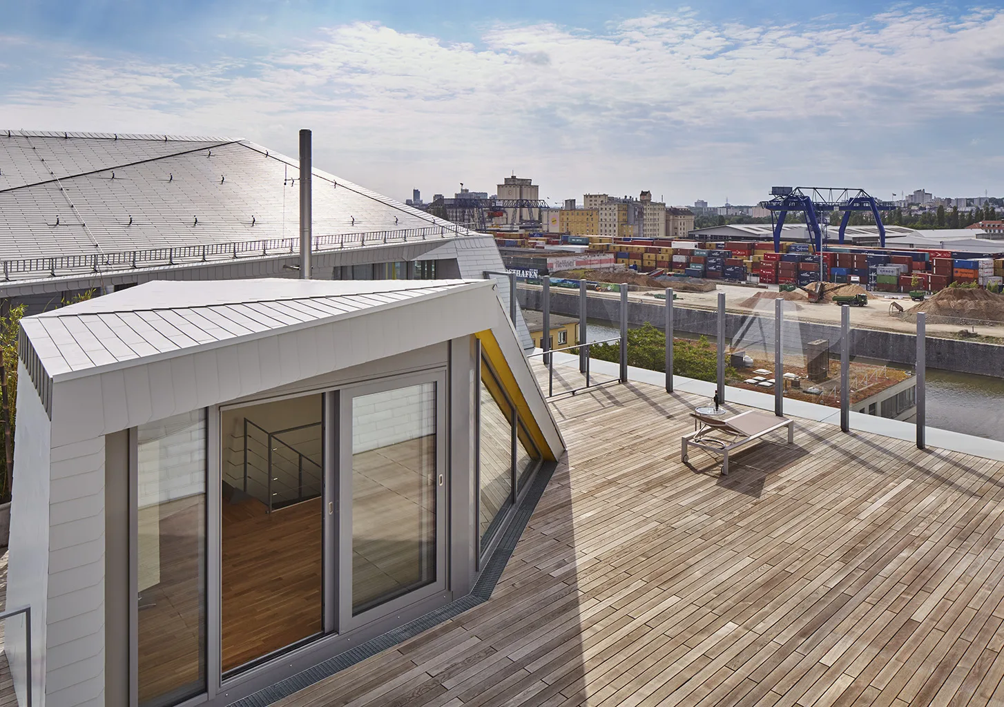Spacious rooftop terrace with glass railing, wooden deck, and angular dormer structure, overlooking city rooftops, cranes, and riverfront industrial landscape.