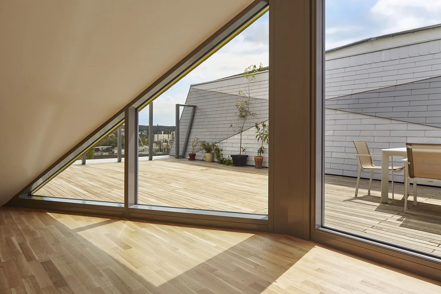 Large triangular window opens to a private rooftop terrace with wood decking, outdoor seating, and expansive city views under a clear sky.