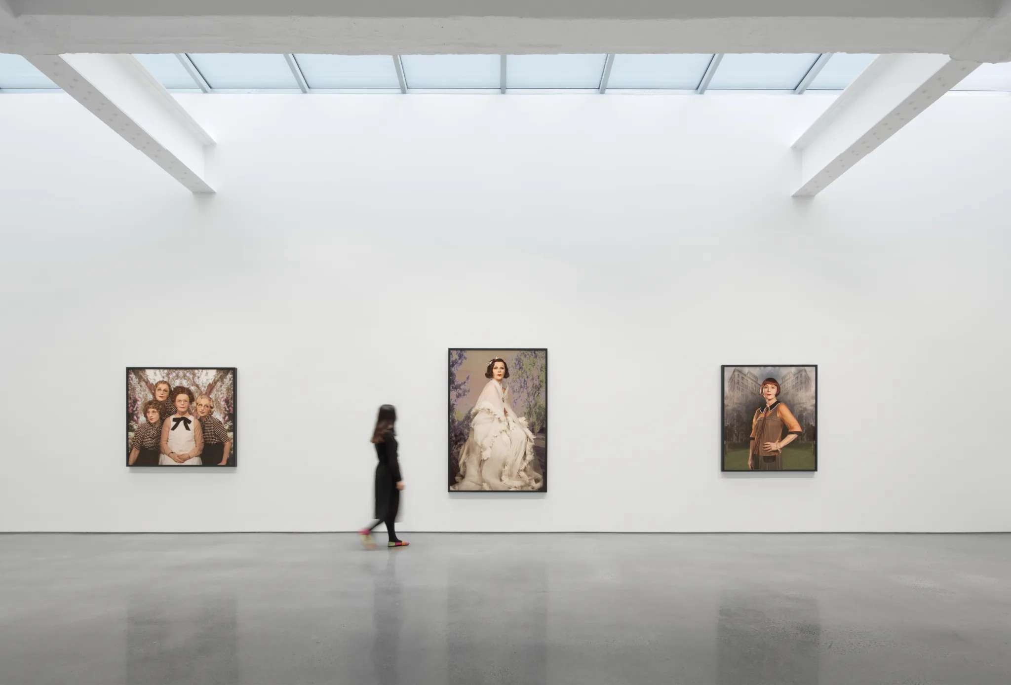 A visitor walks through the Metro Pictures Gallery, featuring large framed portraits displayed on a minimalist white wall under a skylit ceiling.