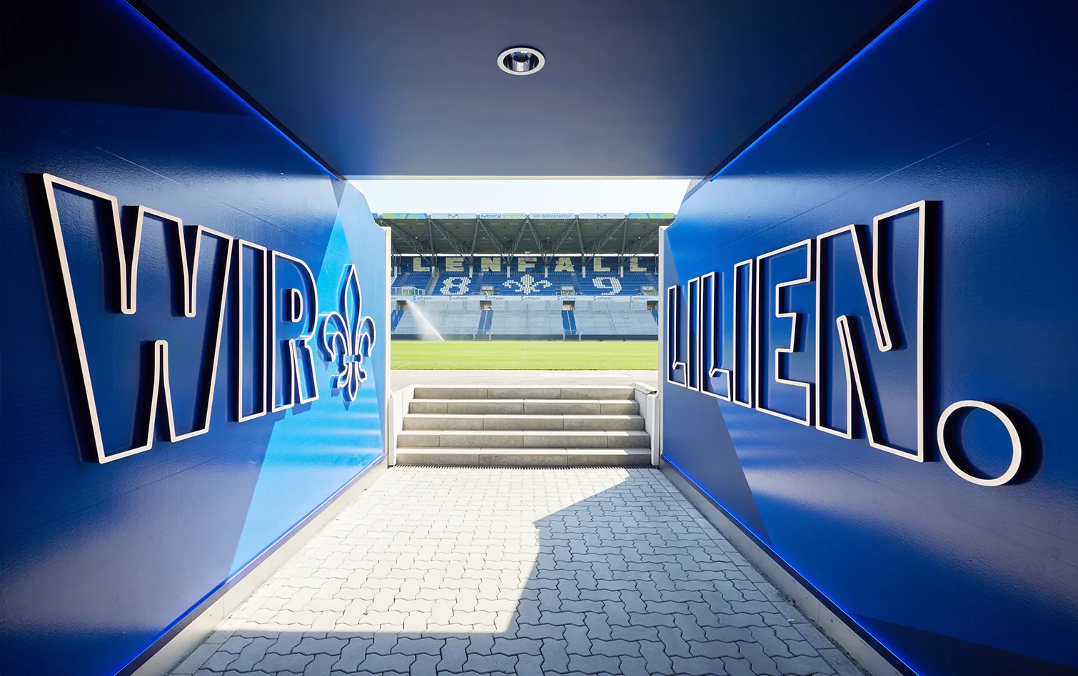 Player tunnel painted in bold blue with “Wir Lilien” text, framing the exit toward the pitch and the stadium seating beyond.