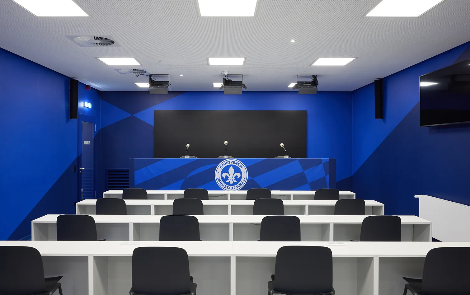 Press conference room with blue walls, team crest backdrop, and rows of white desks with black chairs.
