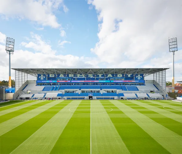View of the renovated Darmstadt football stadium, showing the main grandstand with tiered seating, bold blue accents, and clear sightlines across the freshly striped pitch.
