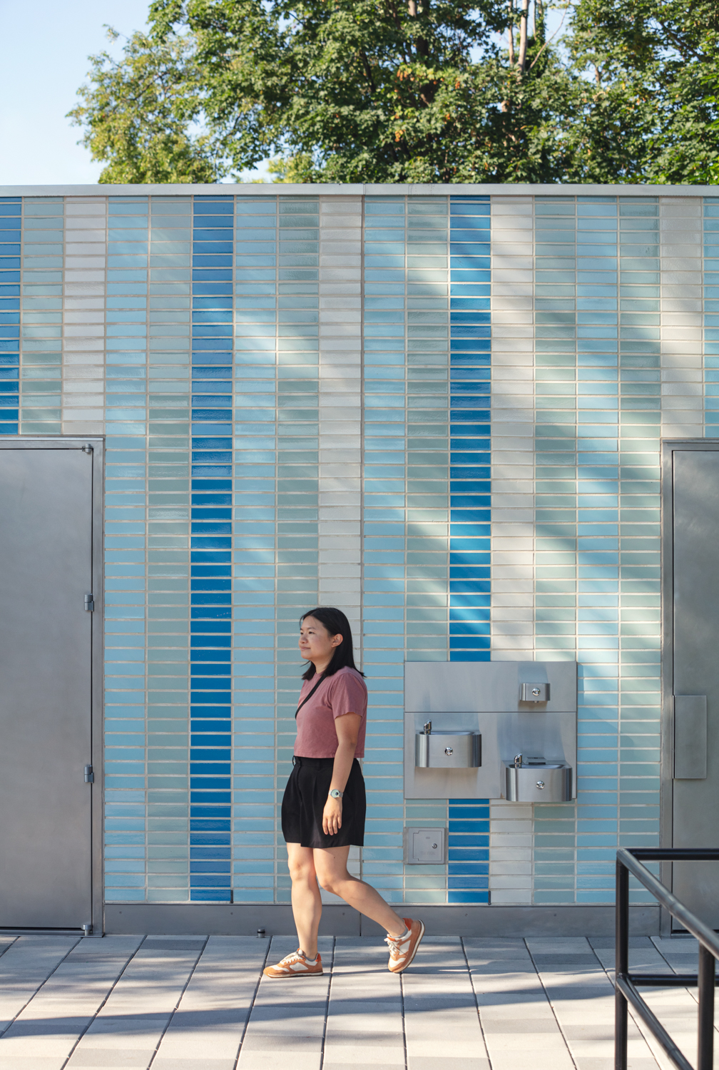 Detail of comfort station façade with vertical blue tile pattern and stainless steel drinking fountains, with a pedestrian walking by.
