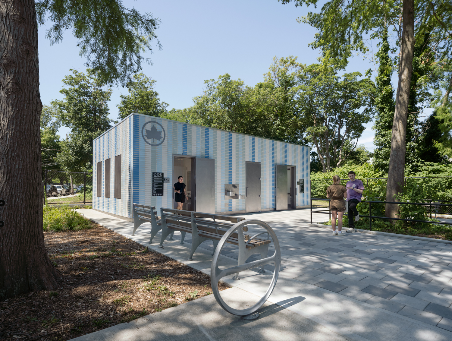 Modern comfort station by 1100 Architect featuring a blue-striped tile façade, integrated seating, and shaded park surroundings designed for accessibility and community use.