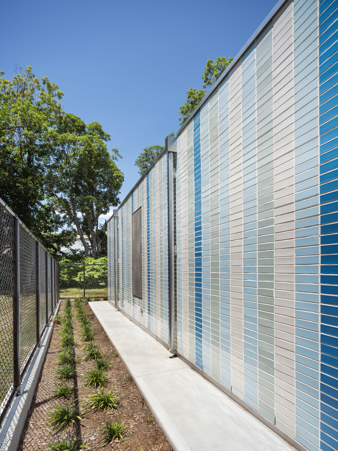 Side view of comfort station with vertical blue and gray tile façade, concrete walkway, and planted landscaping against a bright blue sky.