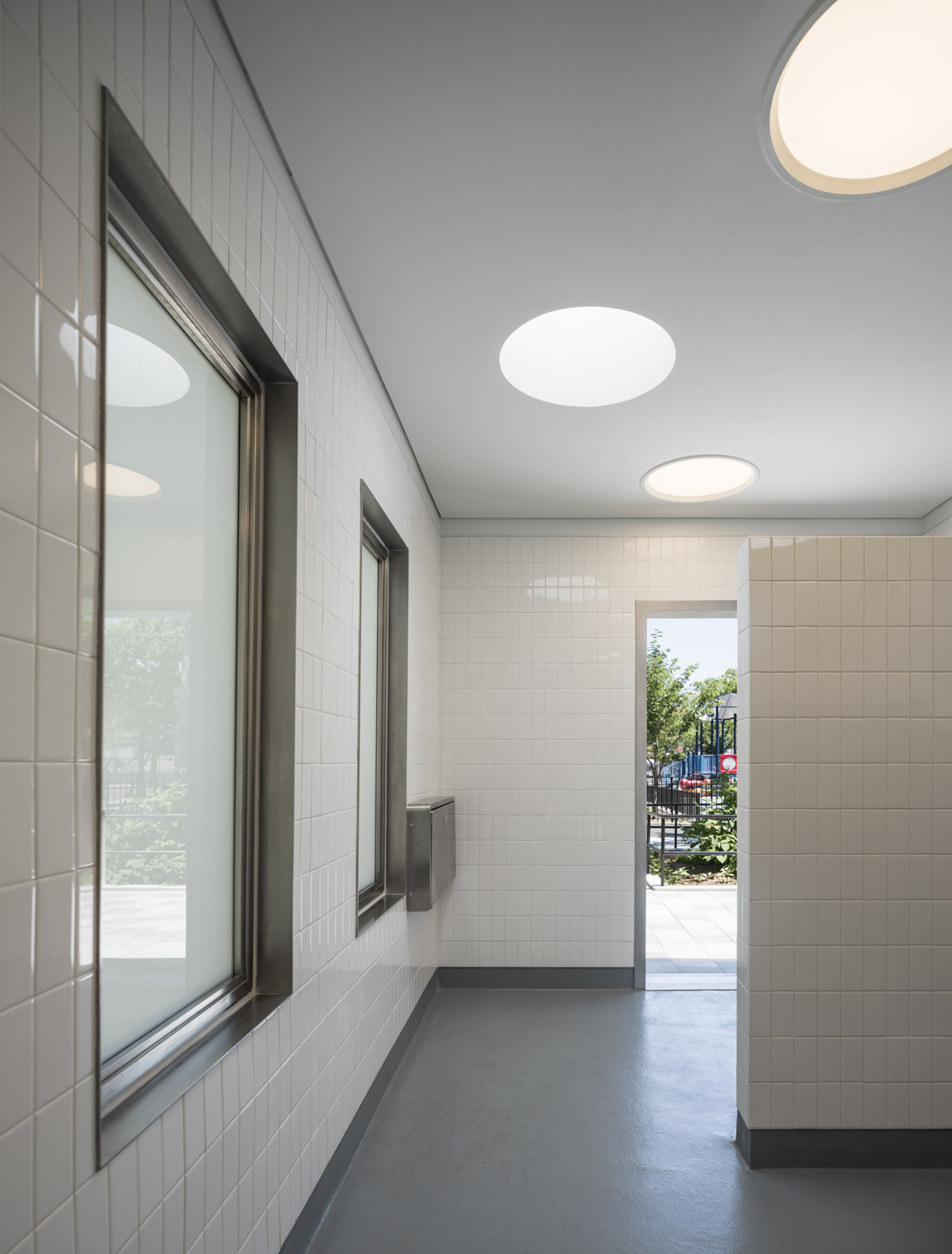 Interior of comfort station with white tiled walls, circular ceiling lights, and stainless steel details, opening out to the playground.