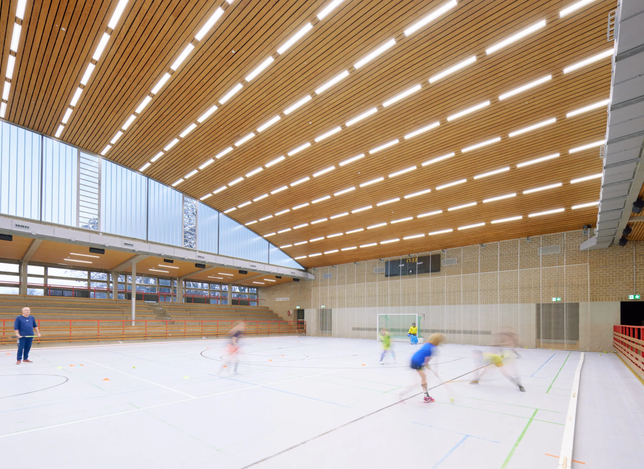 Daylit sports hall with timber-slat ceiling, linear lights, clerestory glazing, and children practicing on the indoor court