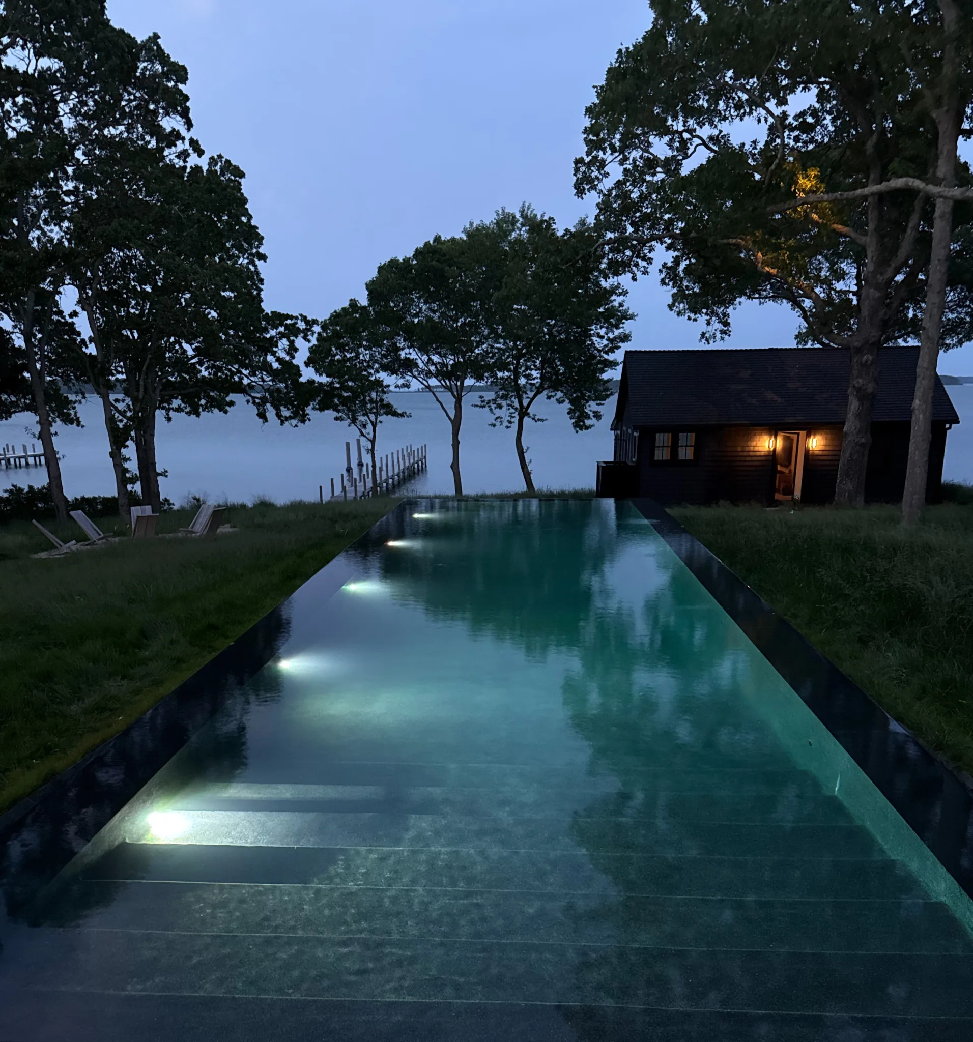 Night view of the illuminated infinity pool at Kelly Klein’s Sag Harbor home by 1100 Architect, overlooking the bay with a black wood cottage glowing in the background.