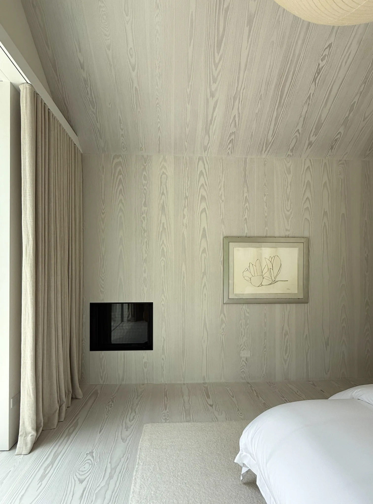 Minimalist bedroom in Kelly Klein’s Sag Harbor house by 1100 Architect, with light wood paneling, soft neutral curtains, framed artwork, and a concealed modern fireplace.