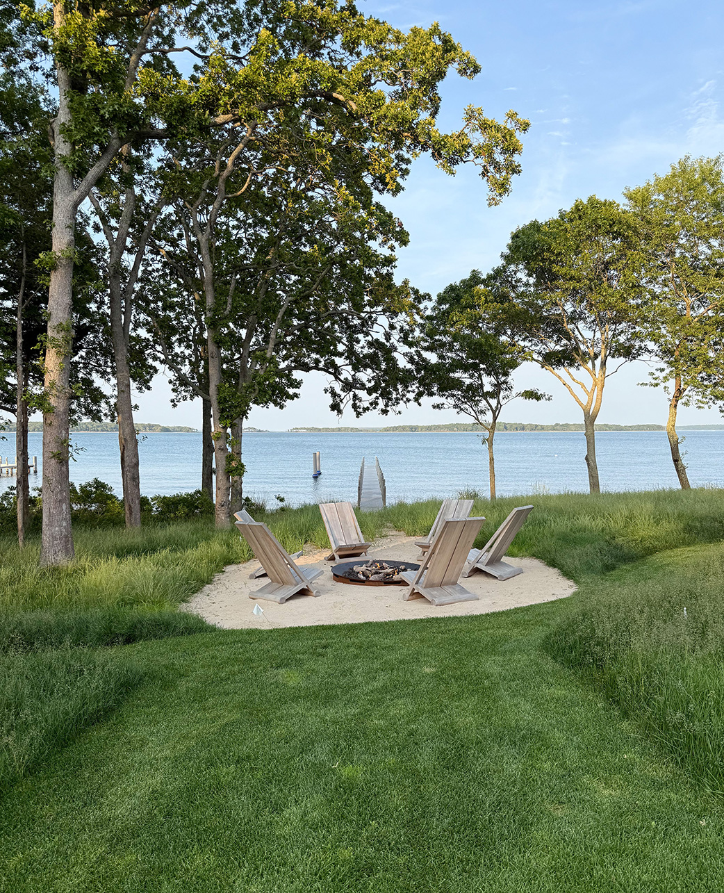 Waterfront fire pit area at Kelly Klein’s Sag Harbor home by 1100 Architect, featuring Adirondack chairs on a sandy circle overlooking the bay through trees.