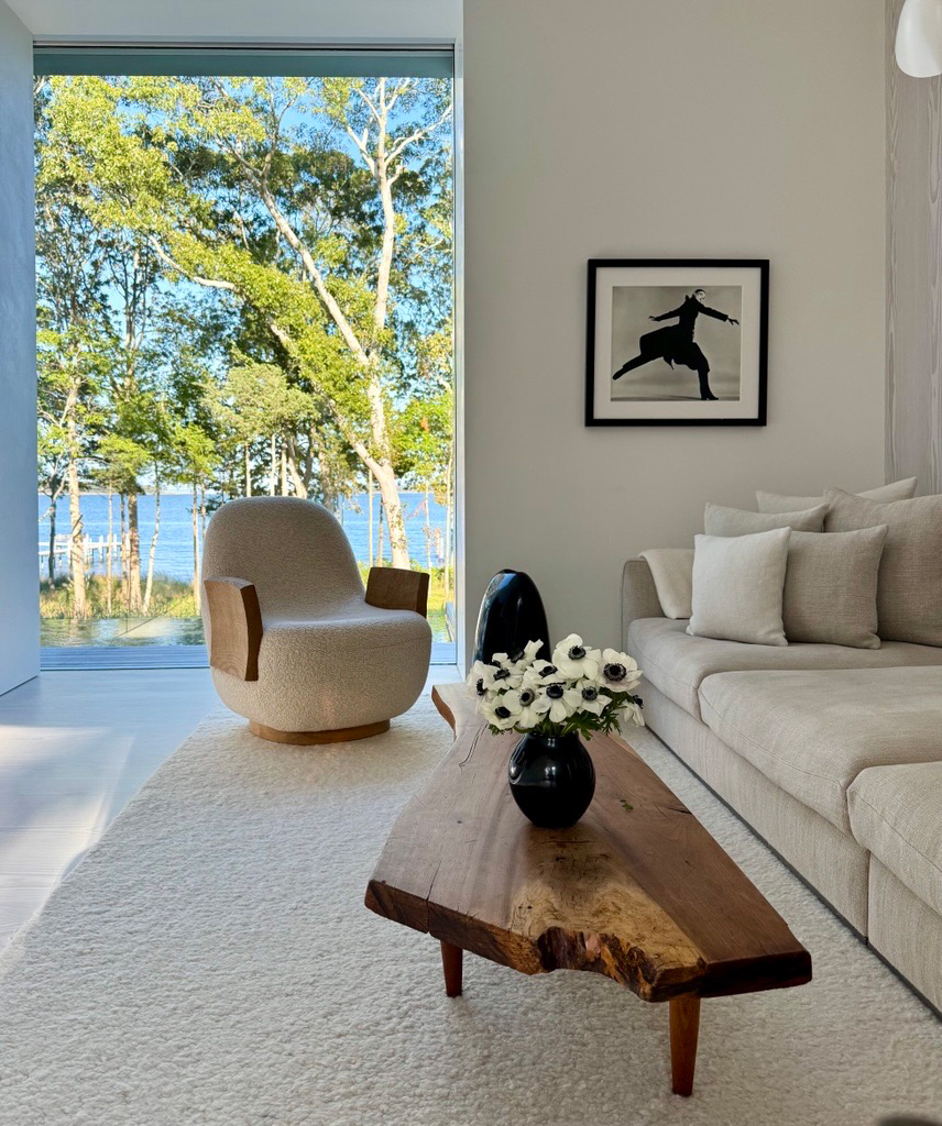 Modern living room with large picture window overlooking Sag Harbor, featuring neutral sofa, sculptural armchair, live-edge wood coffee table, and framed art.