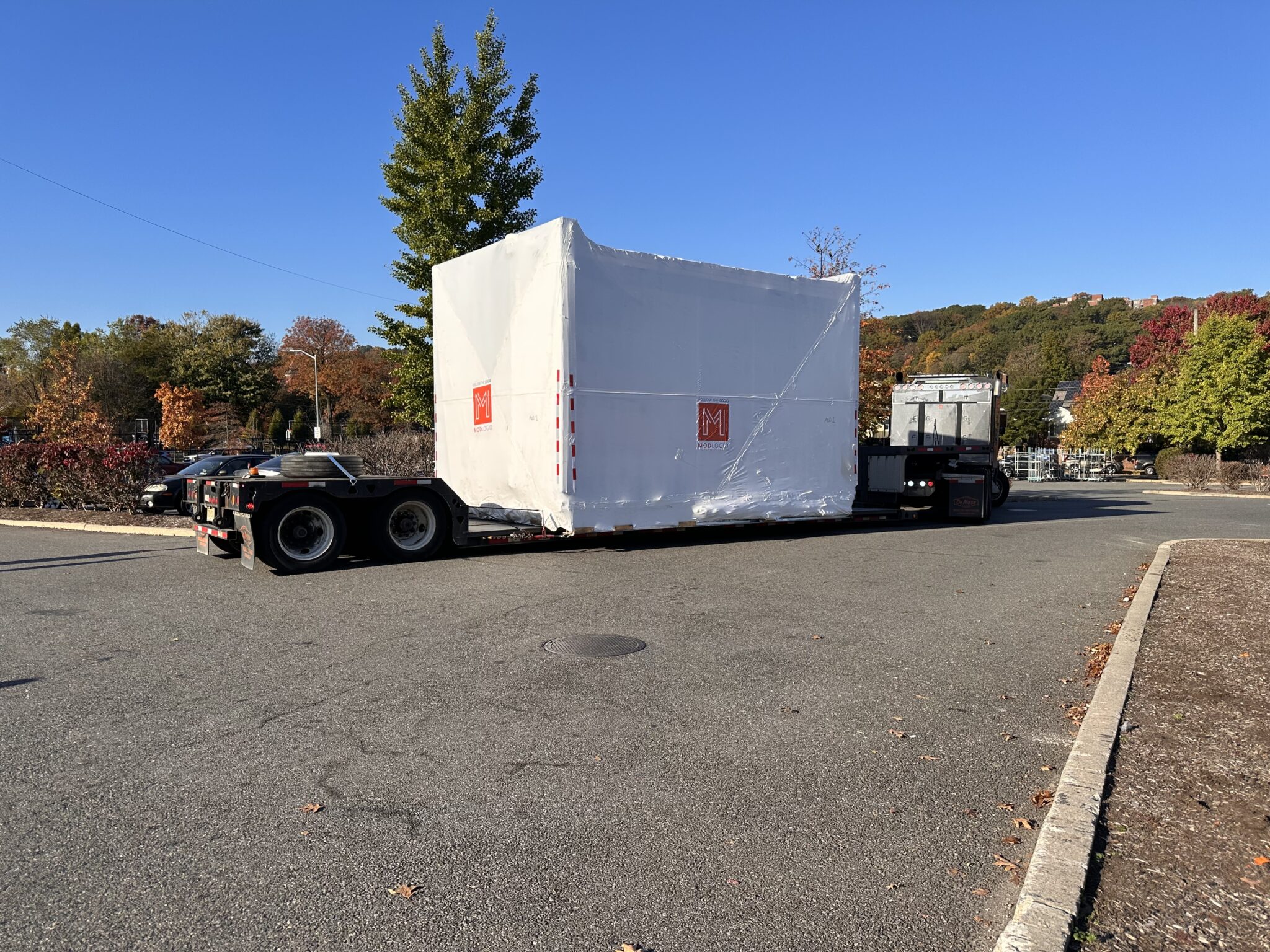 Prefabricated comfort station module wrapped for transport, loaded onto a flatbed truck against a backdrop of autumn trees.