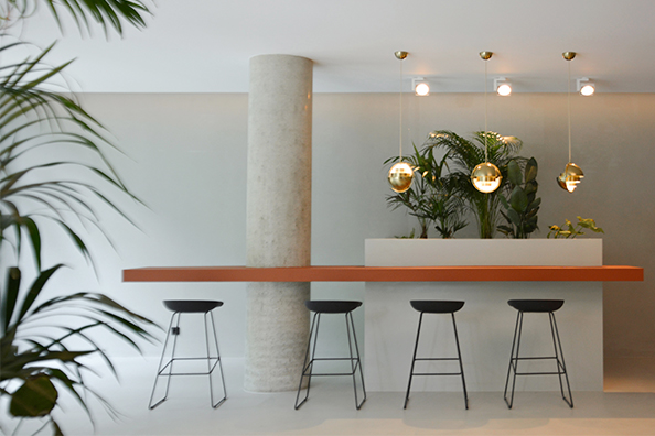 Modernes, helles Foyer eines Bürogebäudes im Frankfurter Stadtzentrum, gestaltet von 1100 Architekten. Im Vordergrund steht ein schmaler, terracottafarbener Hochtisch, begleitet von fünf schwarzen Barhockern mit filigranen Metallbeinen. Über dem Tisch hängen drei kugelförmige Pendelleuchten aus poliertem Messing, die ein warmes, angenehmes Licht erzeugen. Im Hintergrund sind üppige grüne Pflanzen in einem Pflanzbeet integriert, die dem Raum eine natürliche Struktur und Ruhe verleihen. Ein runder Betonsäule unterstreicht den modernen Charakter des Raumes. Die Gestaltung kombiniert natürliche Materialien, klare Linien und eine dezente Farbpalette, die eine einladende und kommunikative Atmosphäre schafft.