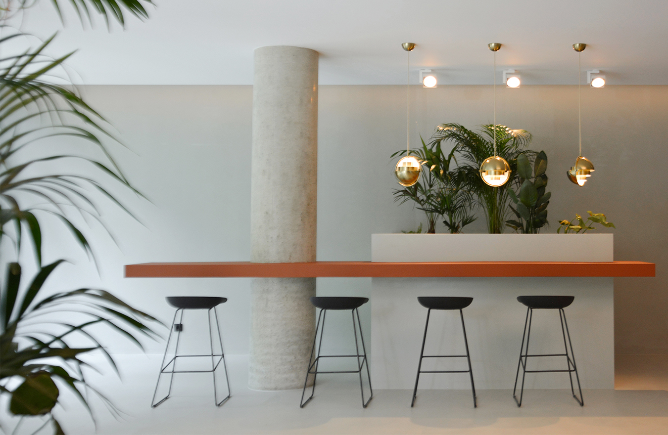 Modernes, helles Foyer eines Bürogebäudes im Frankfurter Stadtzentrum, gestaltet von 1100 Architekten. Im Vordergrund steht ein schmaler, terracottafarbener Hochtisch, begleitet von fünf schwarzen Barhockern mit filigranen Metallbeinen. Über dem Tisch hängen drei kugelförmige Pendelleuchten aus poliertem Messing, die ein warmes, angenehmes Licht erzeugen. Im Hintergrund sind üppige grüne Pflanzen in einem Pflanzbeet integriert, die dem Raum eine natürliche Struktur und Ruhe verleihen. Ein runder Betonsäule unterstreicht den modernen Charakter des Raumes. Die Gestaltung kombiniert natürliche Materialien, klare Linien und eine dezente Farbpalette, die eine einladende und kommunikative Atmosphäre schafft.