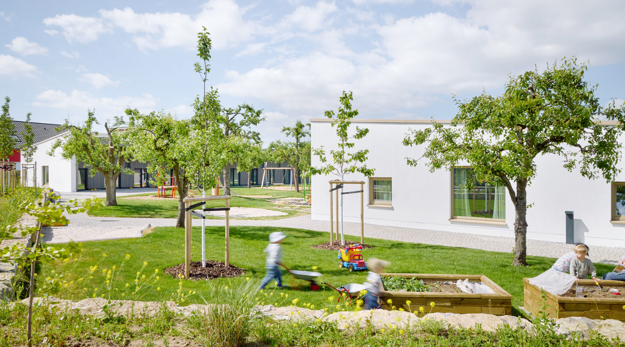 Kindergarten and daycare center in Niddatal with white modern buildings, outdoor play areas, garden beds, and children playing among fruit trees.