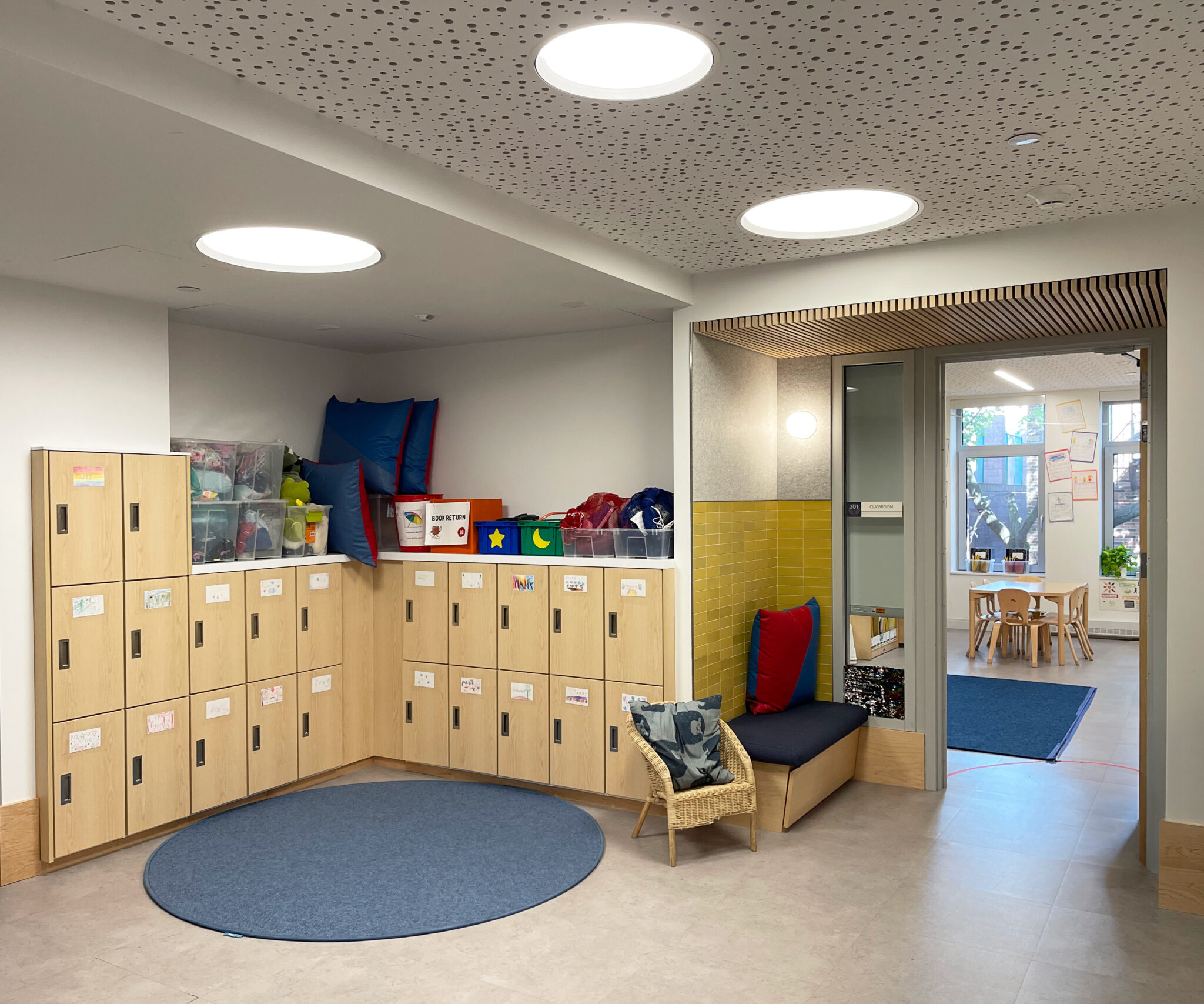 School interior with child-height lockers, reading nook, and adjacent classroom visible through glass doors.
