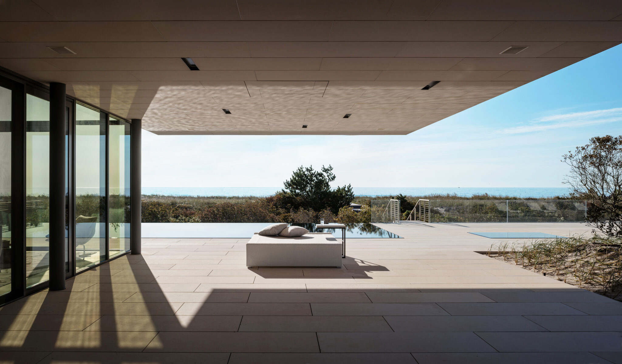 Covered patio with lounge seating and seamless indoor-outdoor connection to the pool and coastal horizon.