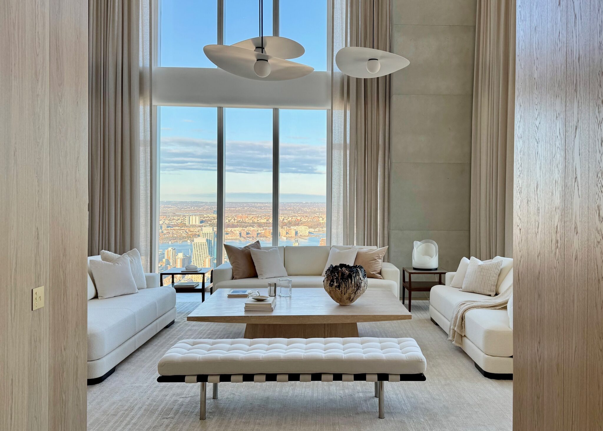 A serene, light-filled penthouse living room featuring two cream sofas, a sculptural coffee table, and panoramic views through floor-to-ceiling windows.