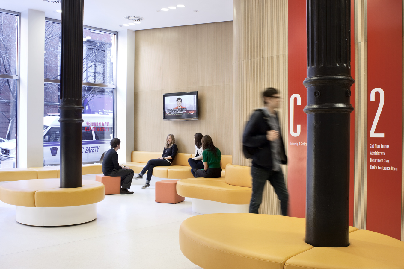 Lobby seating area with circular upholstered benches, a wood-paneled wall, and students gathered around a wall-mounted screen.