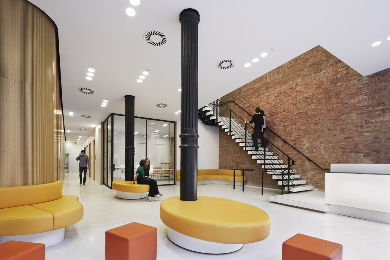 Open lobby with exposed brick, contemporary seating, and a central stair with metal railings leading to the upper level.