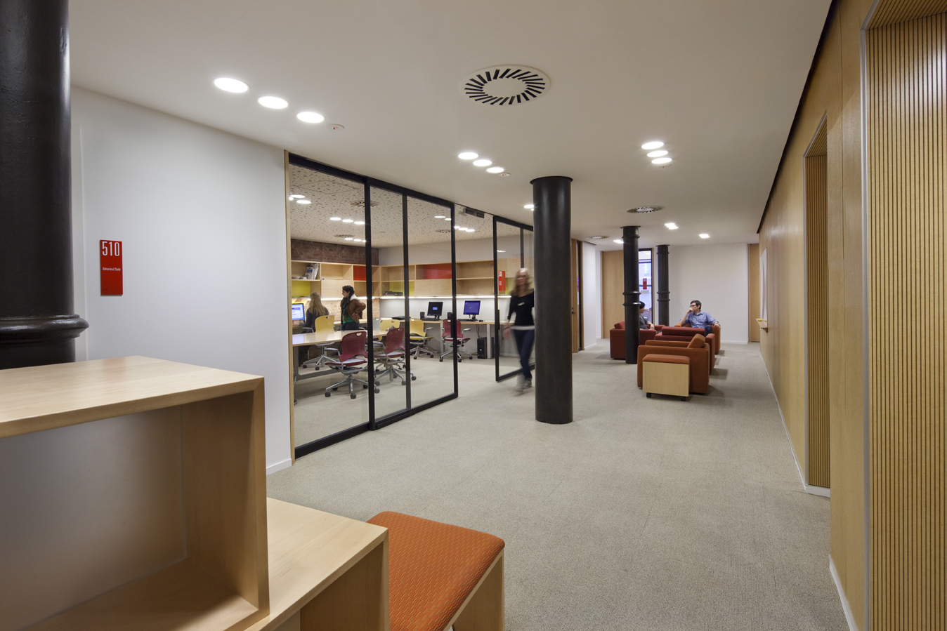Computer lab and study lounge separated by a full-height glass partition, with seating areas arranged along a carpeted circulation path.