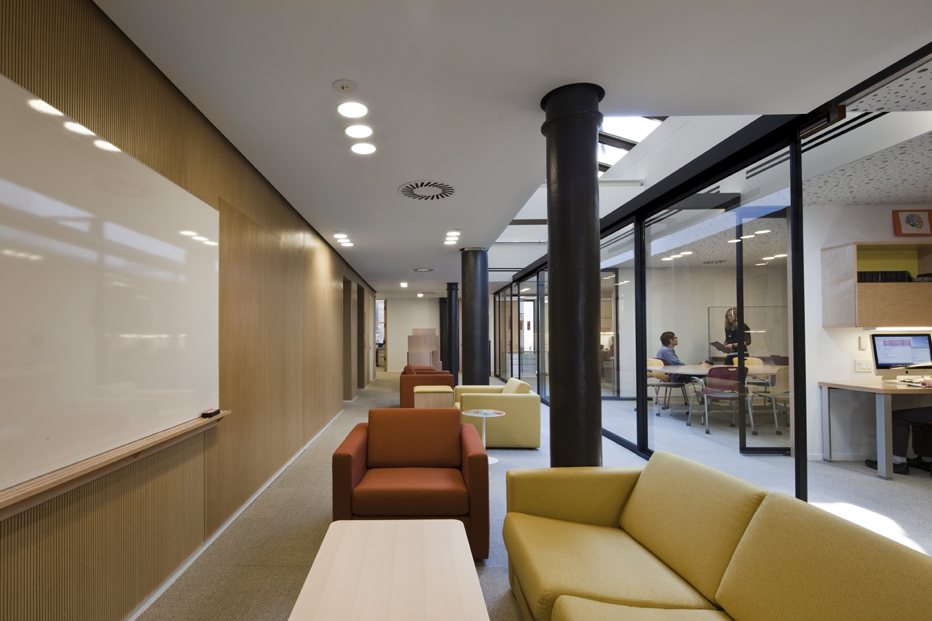 Lounge area with upholstered seating in warm tones, framed by black structural columns and a wood-accented wall, adjacent to glass-walled offices.