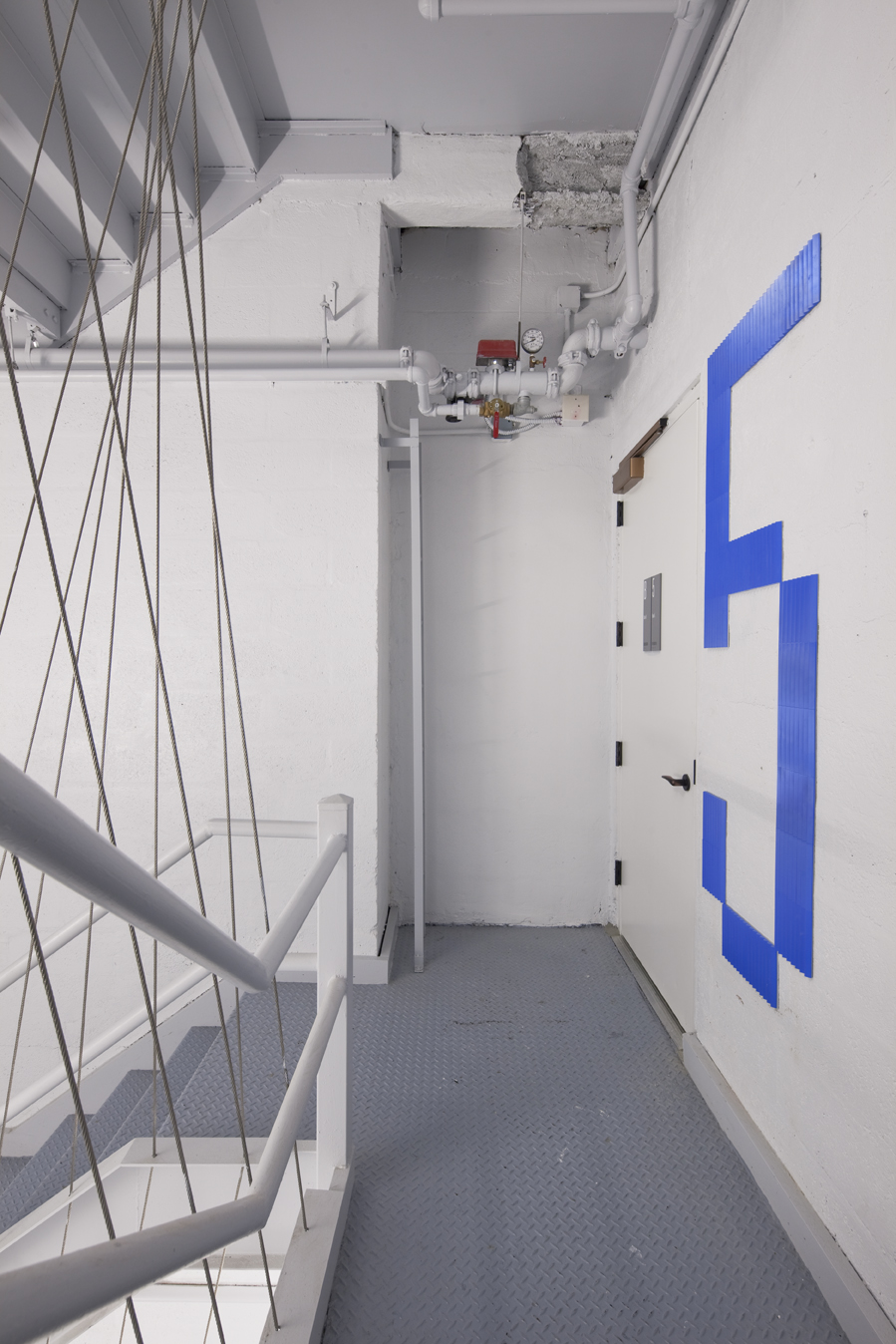 Minimal stair landing with exposed pipes, a white metal railing, and a large blue number five applied to the wall for wayfinding.