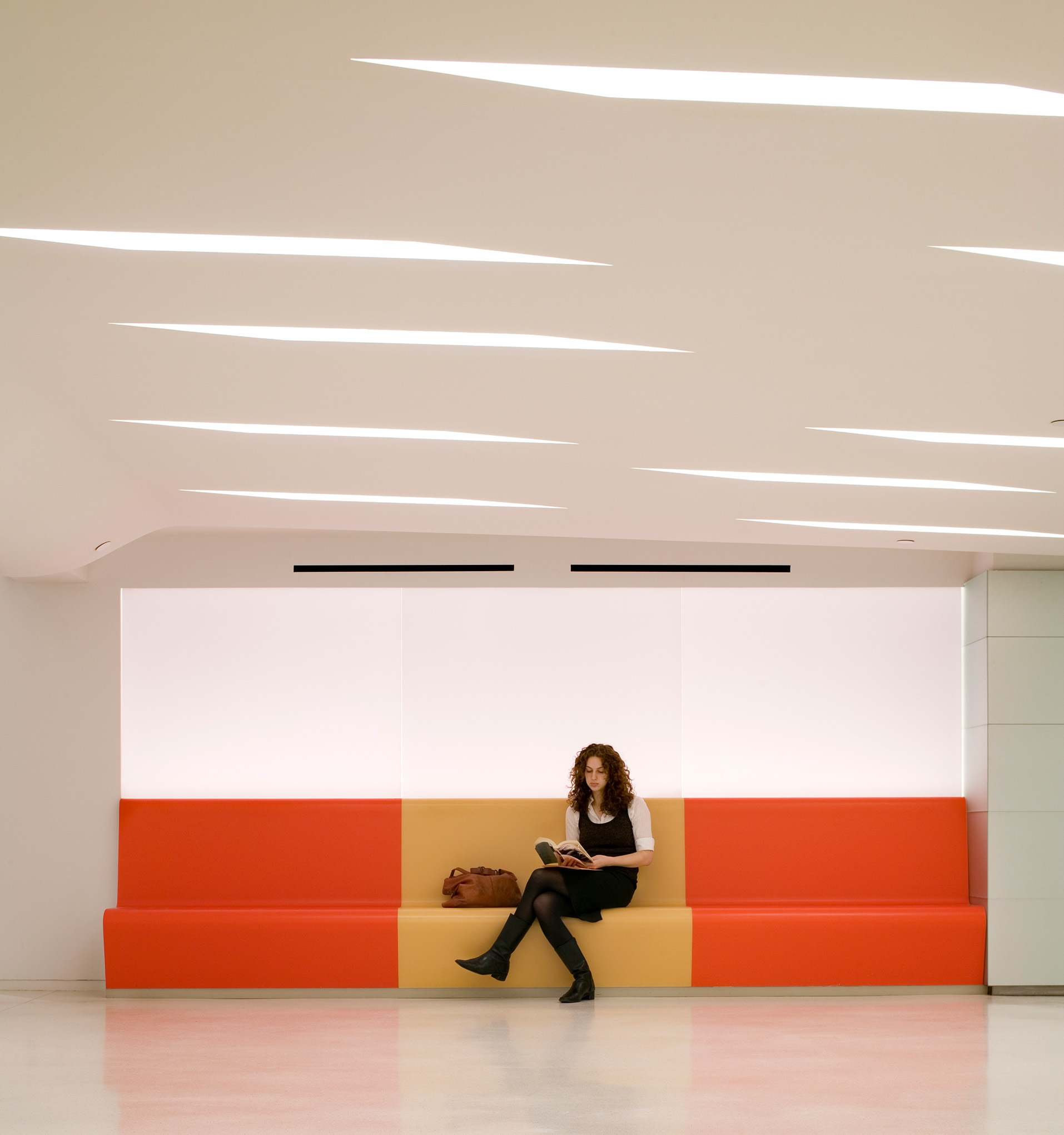 A student sits reading on a long orange-and-tan built-in bench beneath a sculpted ceiling with recessed linear lighting in The New School lobby.
