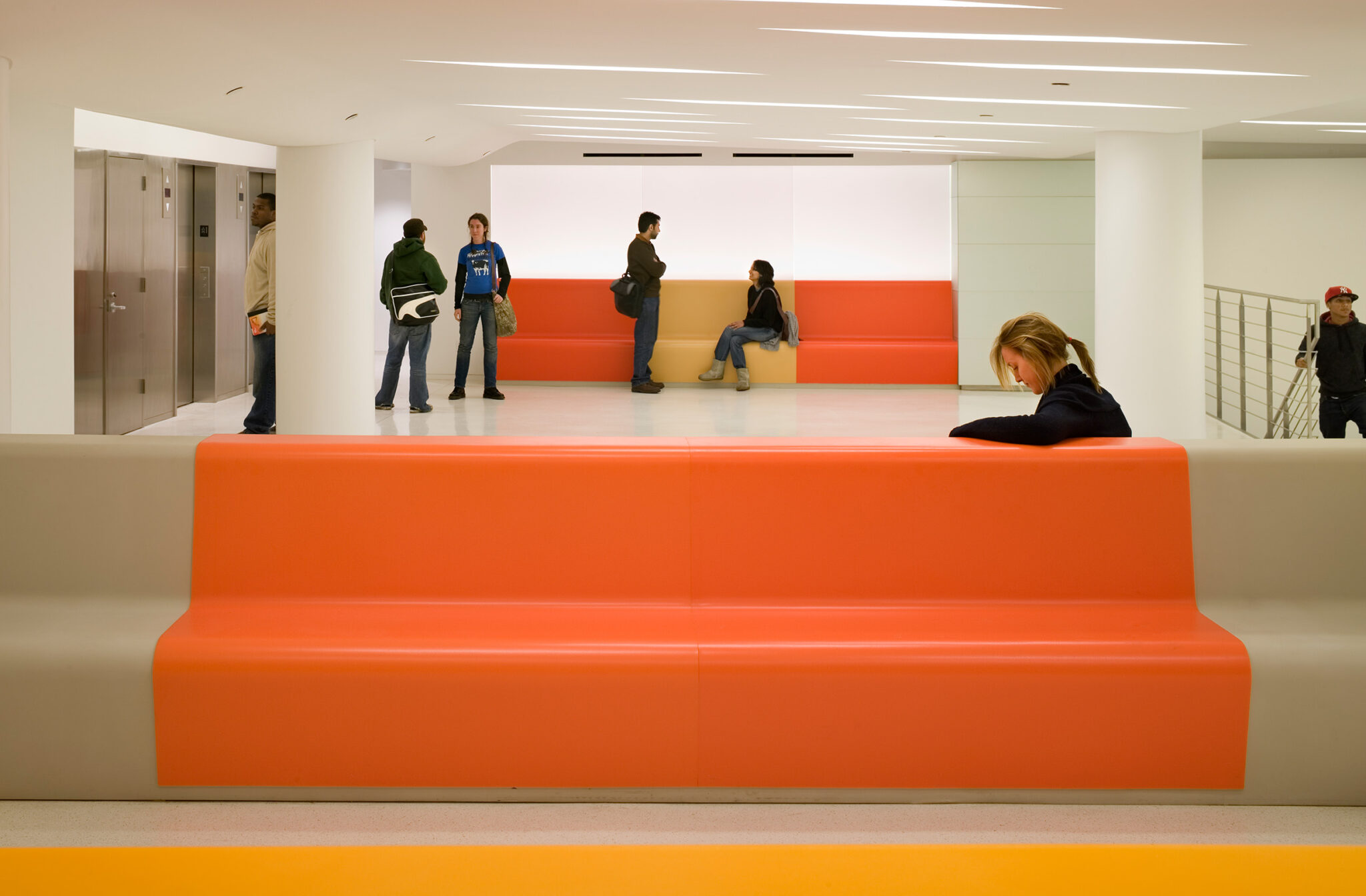 A bright lower-level lounge at The New School featuring integrated orange-and-beige seating, illuminated wall panels, and linear ceiling lights, with a student reading on the bench.