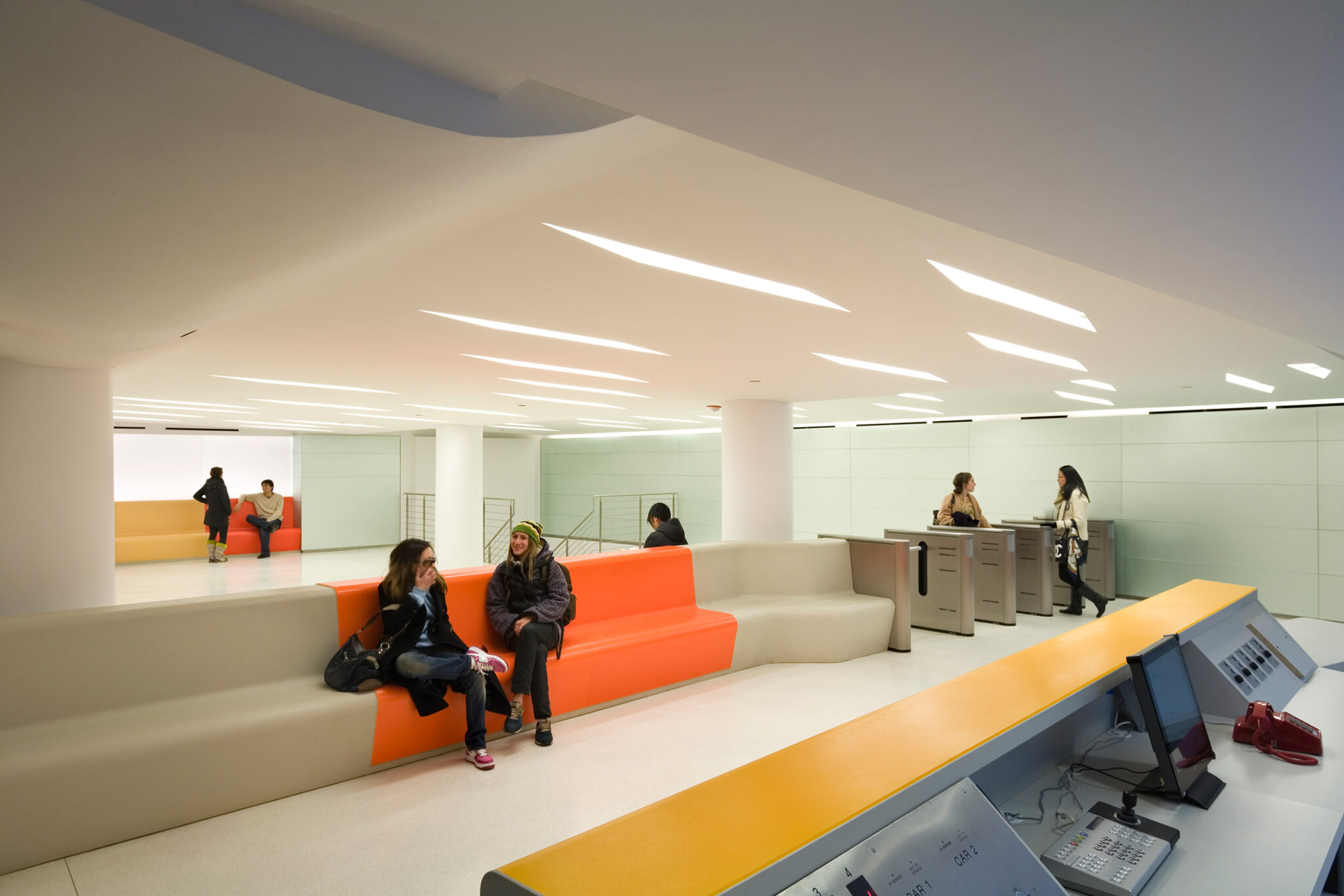 A spacious lobby at The New School with sculpted orange seating, white terrazzo floors, and linear ceiling lighting, where students gather near the elevator core.