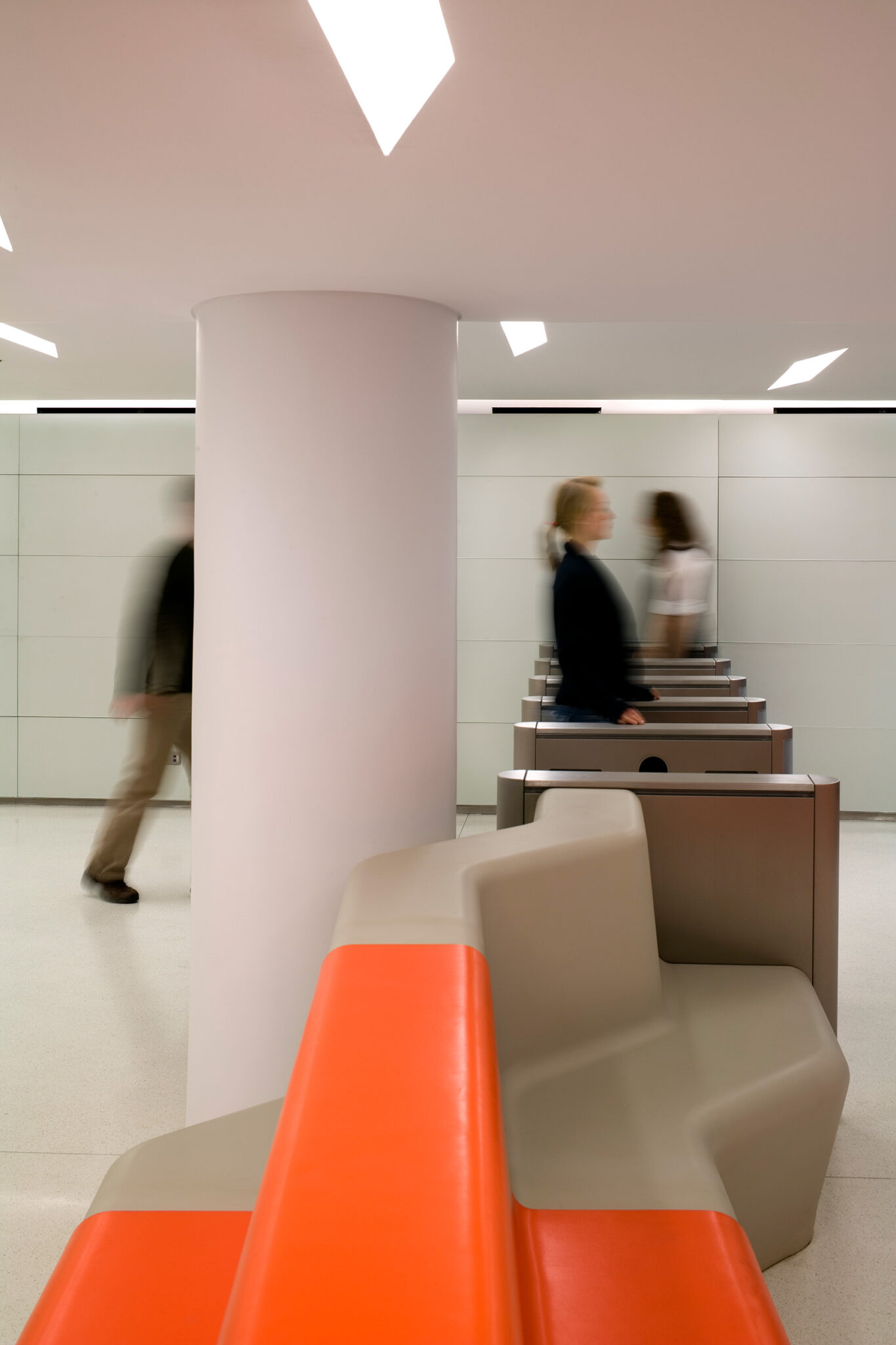 A view from the security desk at The New School showing custom orange and beige lounge seating, stainless-steel turnstiles, and soft, diffused lighting across the open lobby.