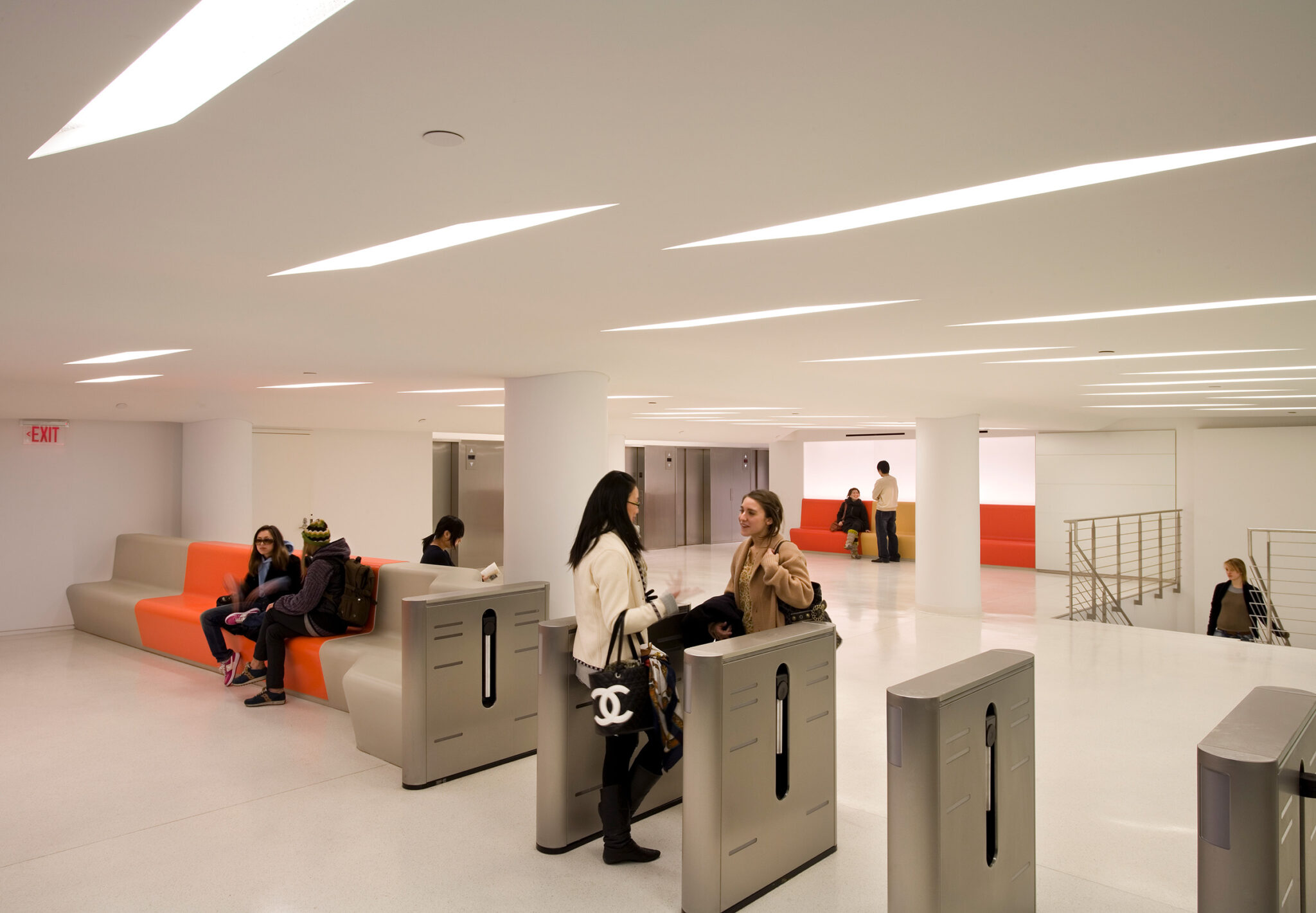 Students move through stainless-steel turnstiles in The New School’s lobby, framed by sculpted lounge seating and a ceiling with angular recessed lighting.