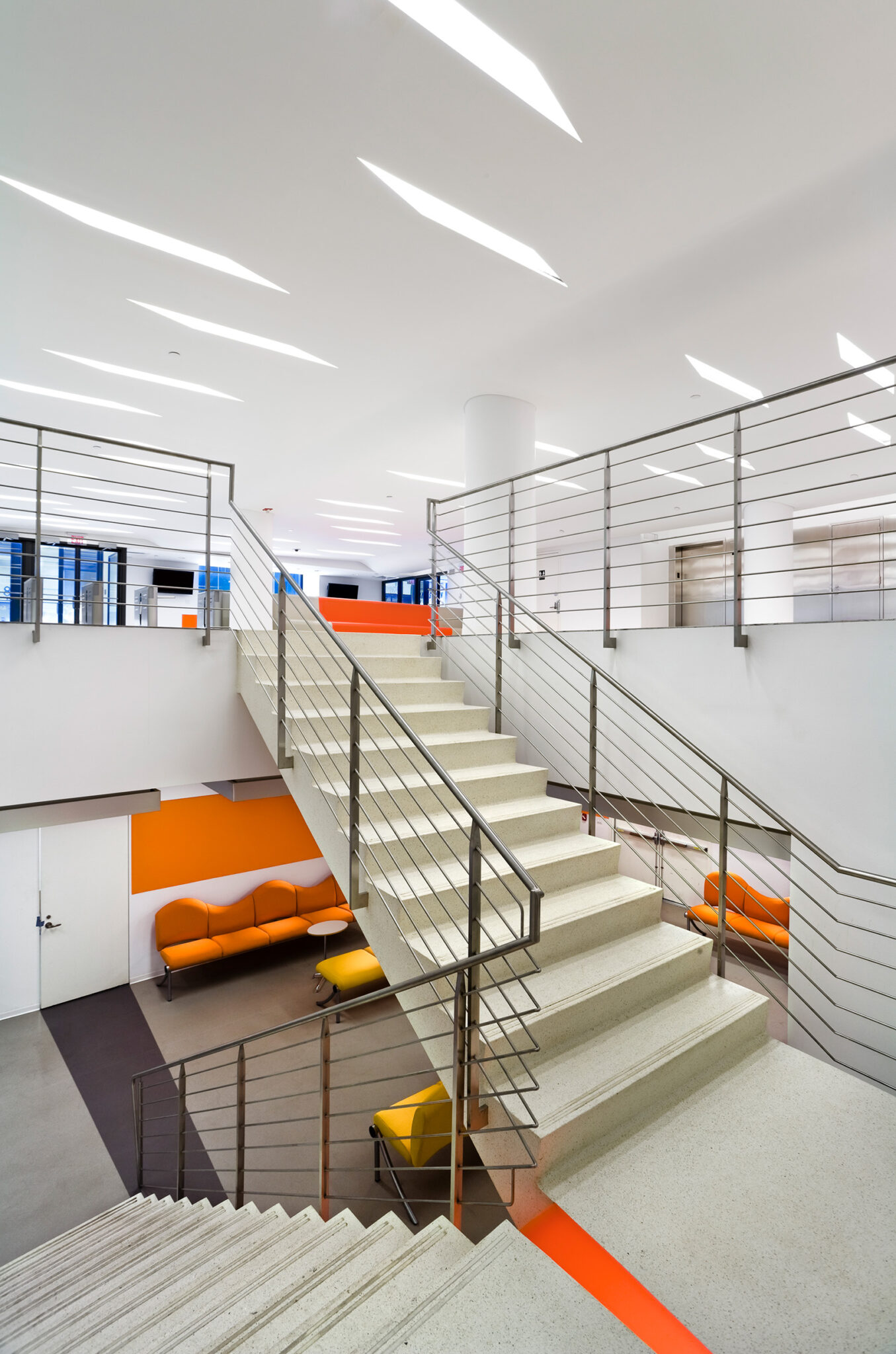 The New School lobby with turnstiles, integrated lounge seating in beige and orange, and a bright, minimal interior illuminated by elongated recessed ceiling lights.