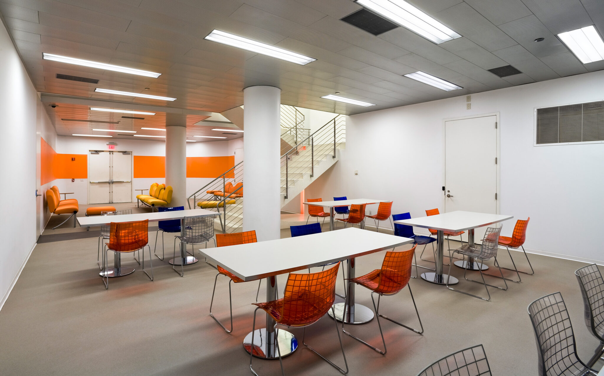 Lower-level communal space at The New School with orange and blue wire chairs, white tables, and column-lined ceilings beneath the central stair.