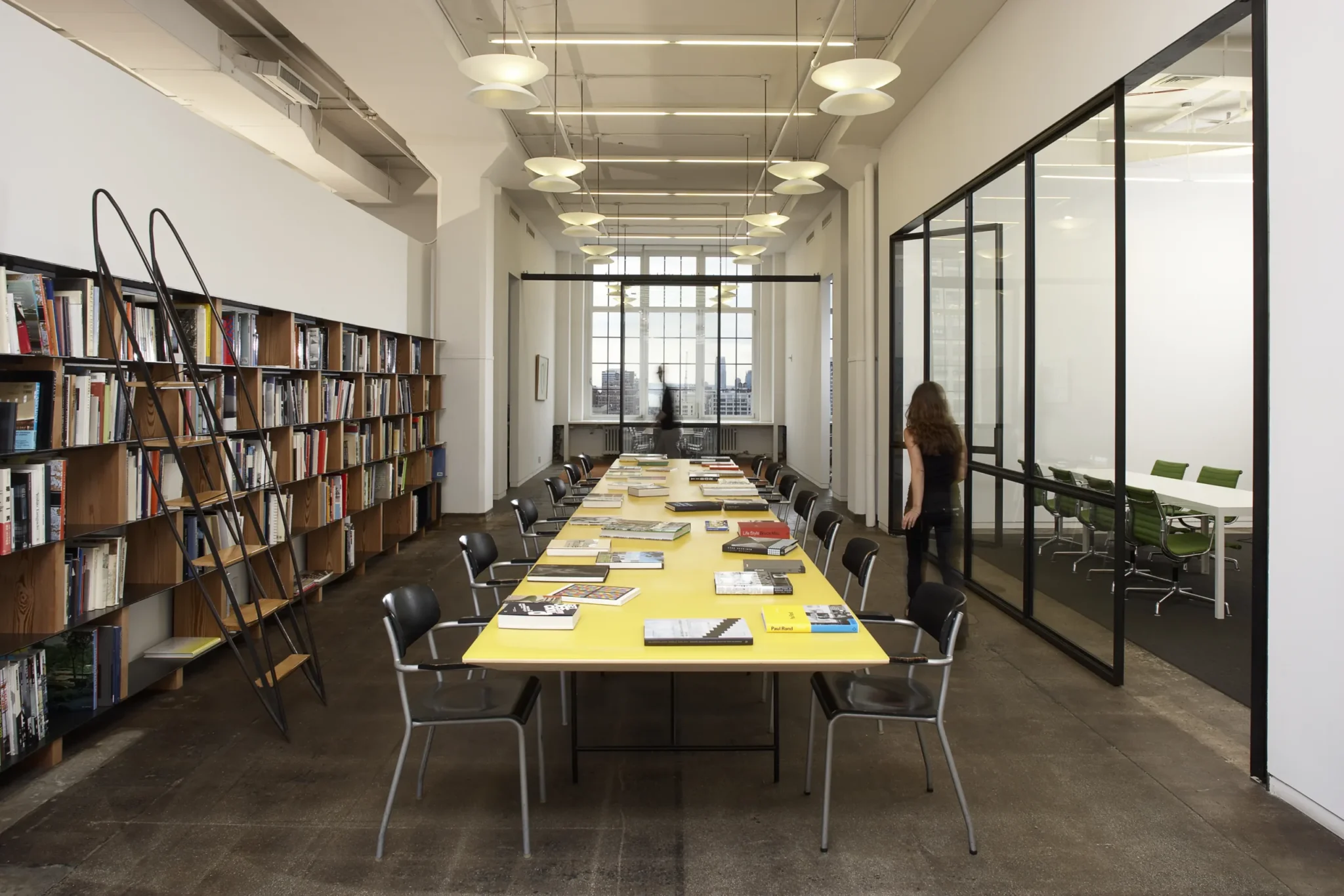Blick in einen hellen Konferenzbereich unseres Büros in New York City: Ein langer gelber Tisch mit ausliegenden Büchern steht zwischen schwarzen Stühlen; links sind hohe Bücherregale, rechts ein verglaster Besprechungsraum. Am Ende des Raums fällt Tageslicht durch große Fenster mit Blick auf die Skyline.