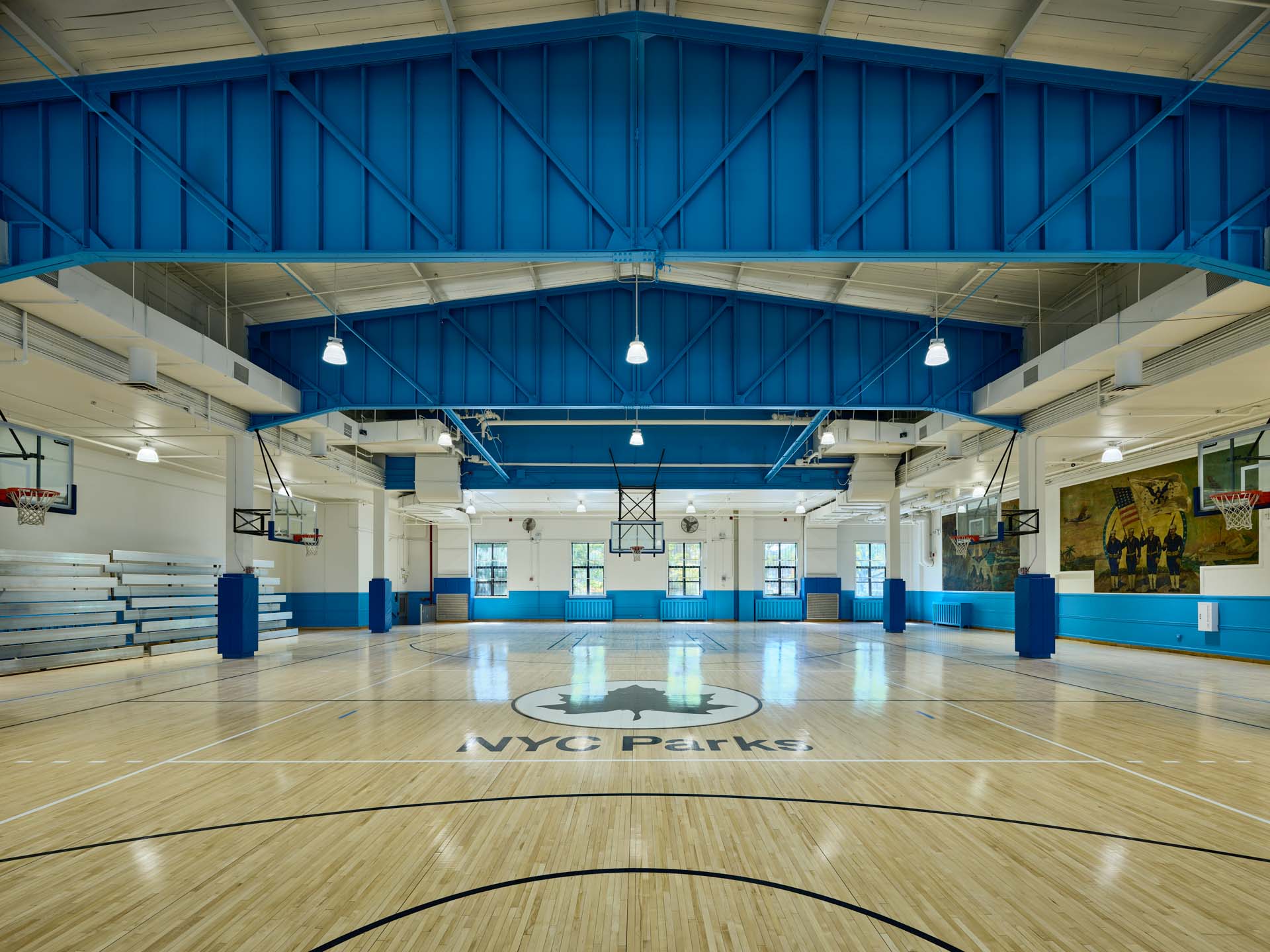 Renovated NYC Parks recreation center basketball court with blue painted steel roof trusses and wood floor.
