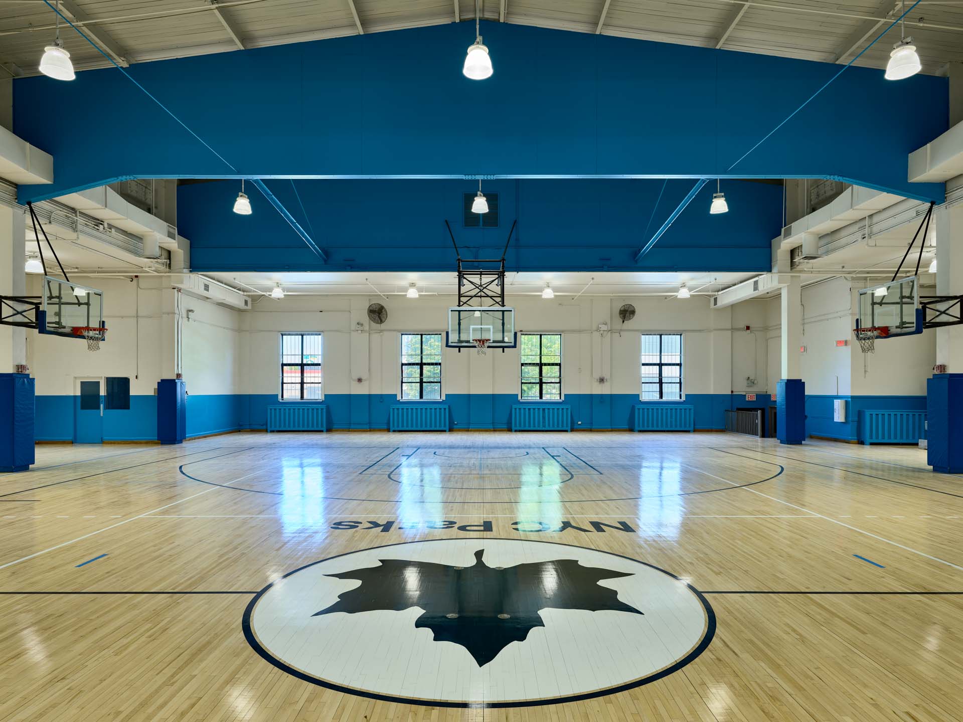 Full-court view of restored NYC Parks indoor basketball gymnasium with blue structural steel trusses and bleacher seating.