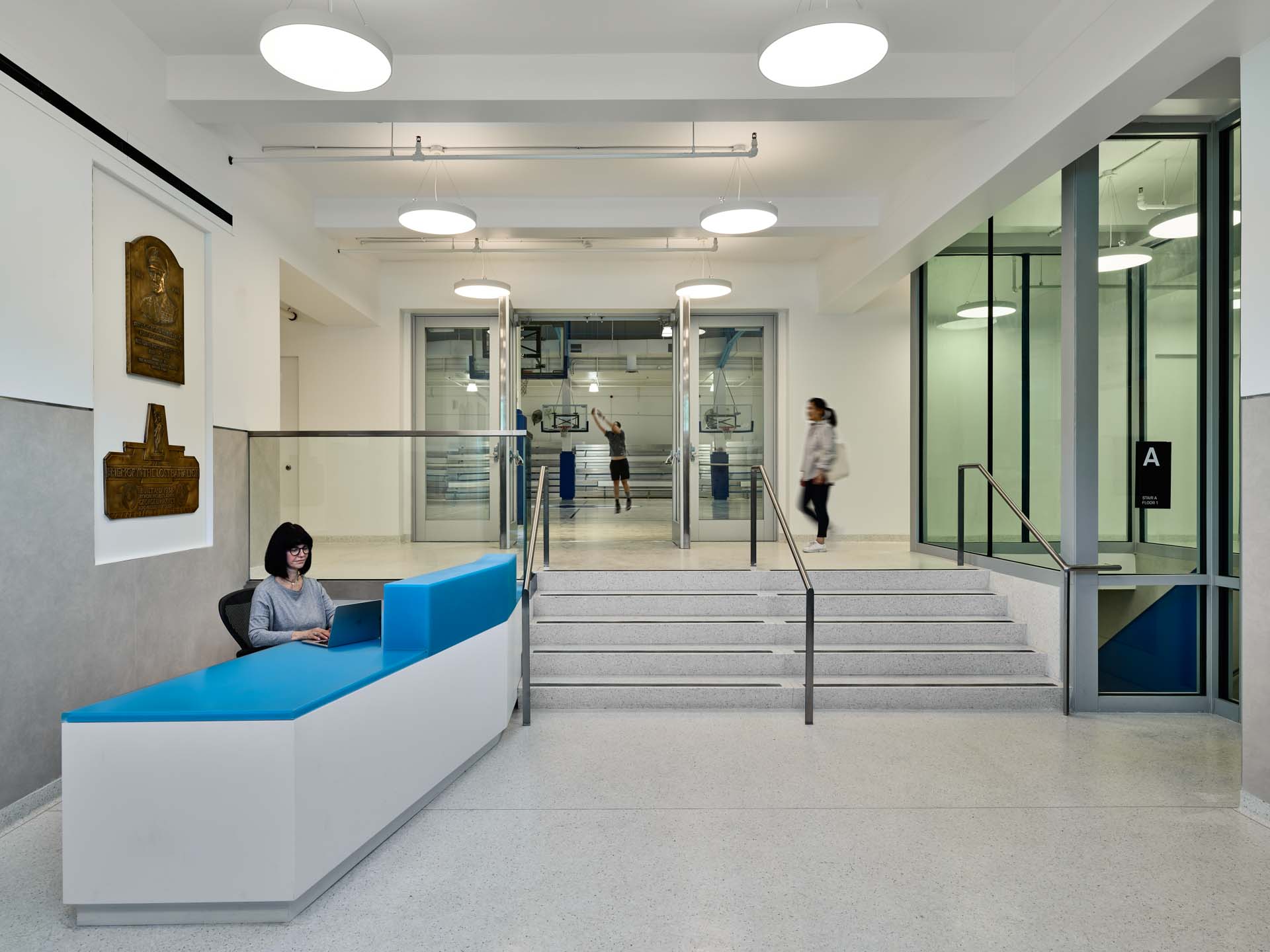 Renovated public recreation center lobby with blue reception desk, terrazzo floors, and glazed views to basketball court.