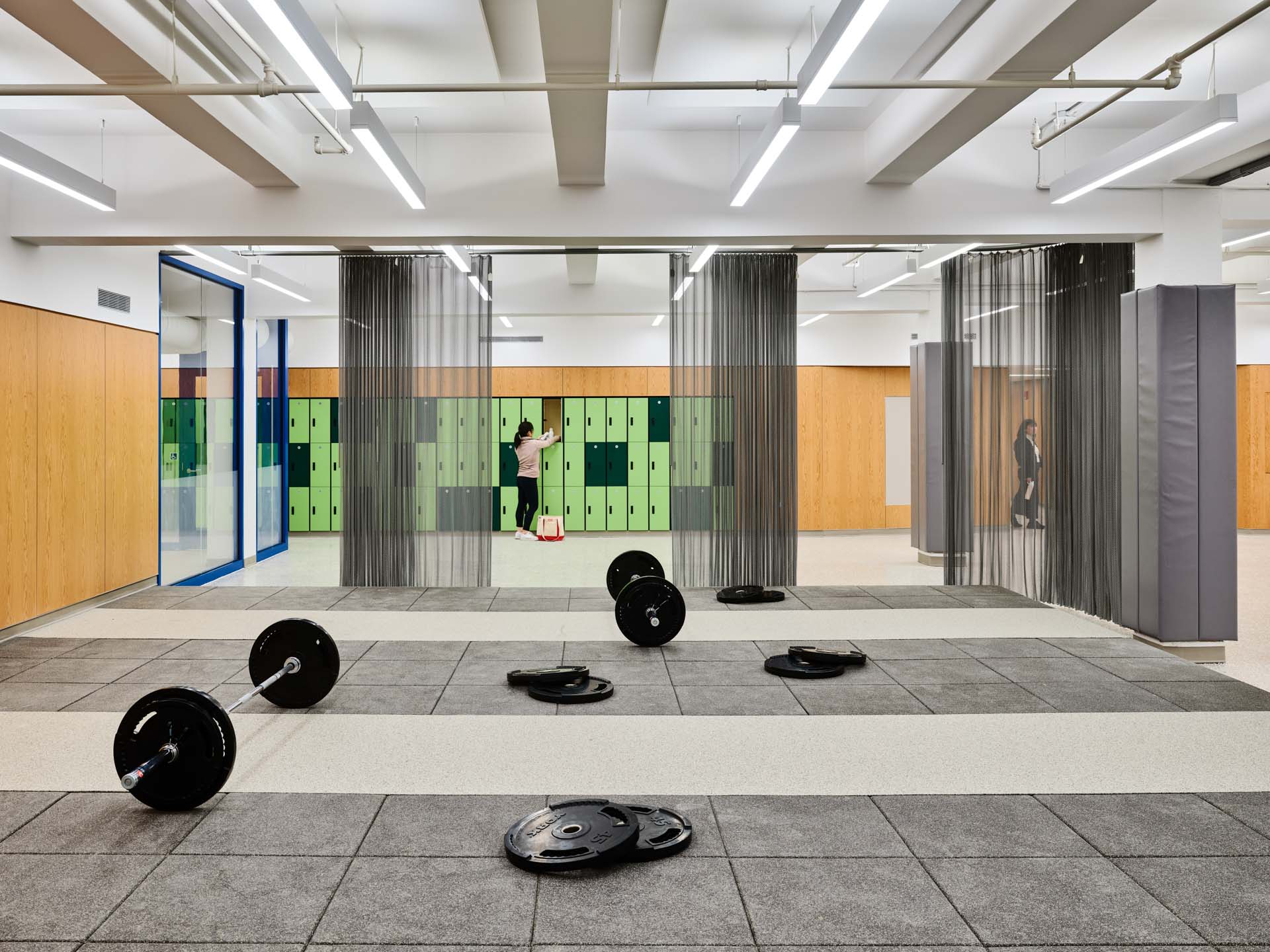 Weight training area with rubber floor, free weights, and locker room beyond in renovated NYC Parks community fitness center.